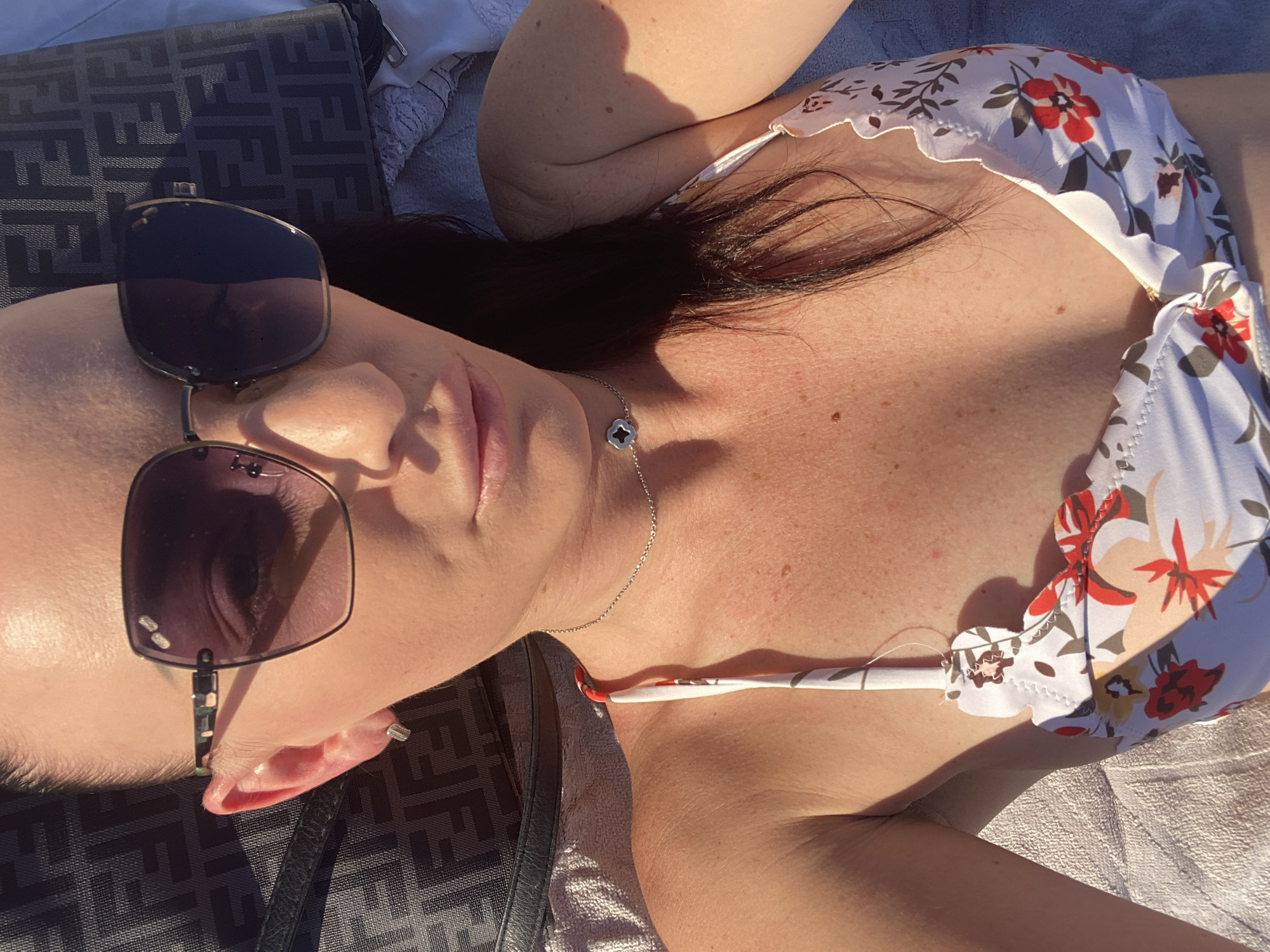 The image shows a person lying down, possibly on a beach or by a pool, given the presence of a towel and a Fendi bag. The person is wearing sunglasses and a floral bikini. The sunlight suggests it's daytime, likely warm weather given the attire. A shadow is cast across the person's chest, indicating the sun's position. The overall mood seems relaxed and summery. The text "FENDI" is visible on the bag.
