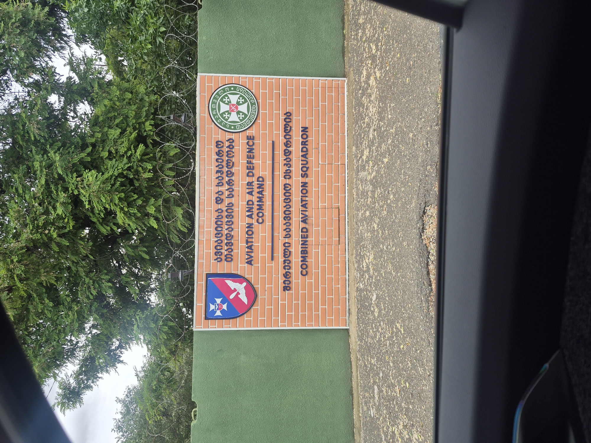 The image shows a sign in English and Georgian that reads "Aviation and Air Defence Command" and "Combined Aviation Squadron." The sign is constructed of brick. A barbed wire fence and green foliage are adjacent to the sign. The image was likely taken in the city of Tbilisi, Georgia.