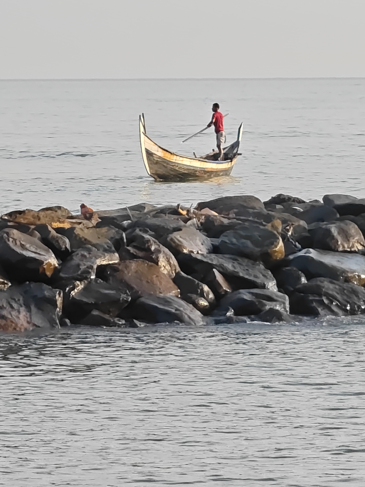 A person is standing in a small boat and rowing. The boat is in the water. In the foreground, there is a rocky breakwater. The sky is overcast. The scene is likely a fishing harbor or coastline in Lhokseumawe, Indonesia.