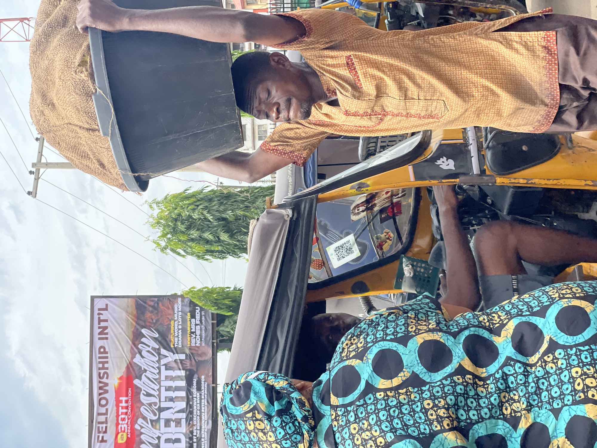 The image shows a man unloading a black bucket filled with a burlap sack into the passenger seat of a yellow tricycle taxi, likely in Onitsha, Nigeria. He is wearing a patterned, long-sleeved shirt and dark trousers. The inside of the taxi is visible, with a passenger in the back wearing a turquoise, yellow, and black patterned fabric. In the background, there is a large billboard advertising "Fellowship Int'l 30th Annual Convention." A pole with power lines and trees are also visible.
