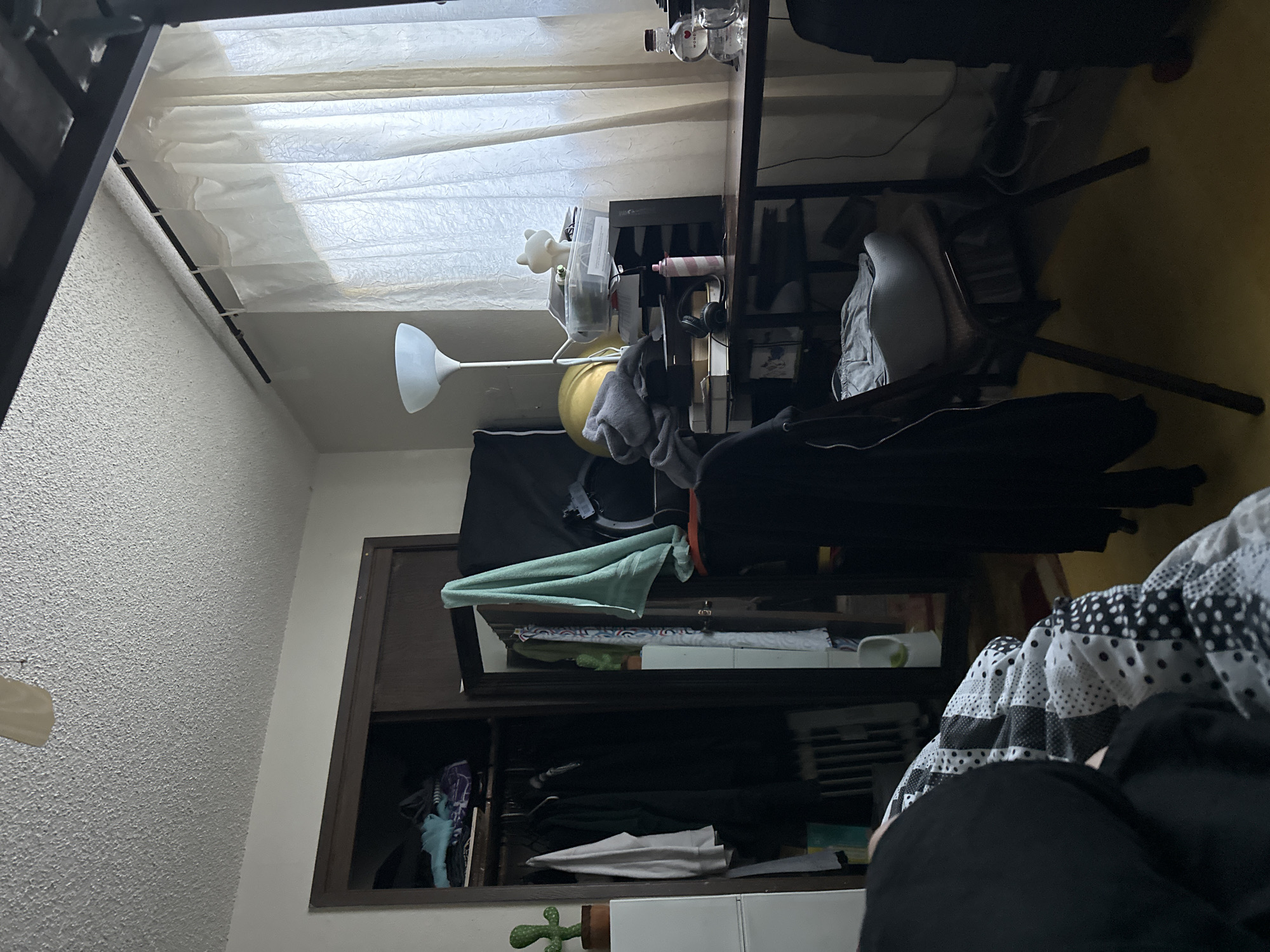 The image depicts a cluttered bedroom interior. A closet filled with clothing is visible on the left side of the frame. To the right, a desk is laden with various items, including a lamp, storage containers, a pink bottle, headphones, and a small white cat figurine. A chair with a light-colored cushion is positioned at the desk. A large window covered by sheer curtains is situated behind the desk. The flooring appears to be a yellow carpet. The scene is dimly lit, and the overall impression is one of disarray. The image was taken in Vancouver, United States of America.