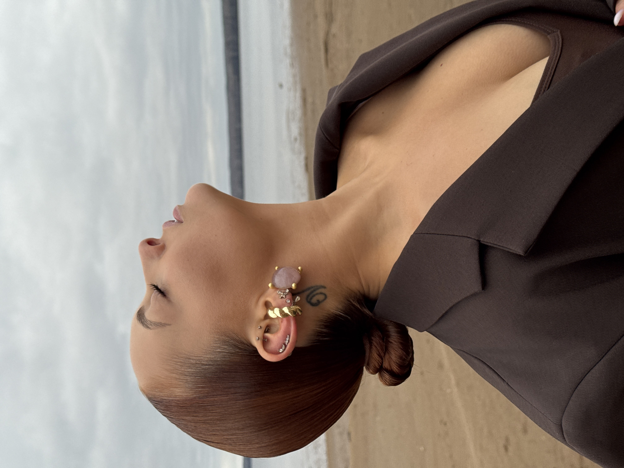 A woman with her head tilted back is standing on a beach. She has brown hair pulled back in a bun and is wearing a brown jacket. Her right ear is adorned with multiple piercings and earrings. The beach and overcast sky are visible in the background. While not explicitly confirmed in the image, the question prompt implies that the location is Chaikovsky, Russia.