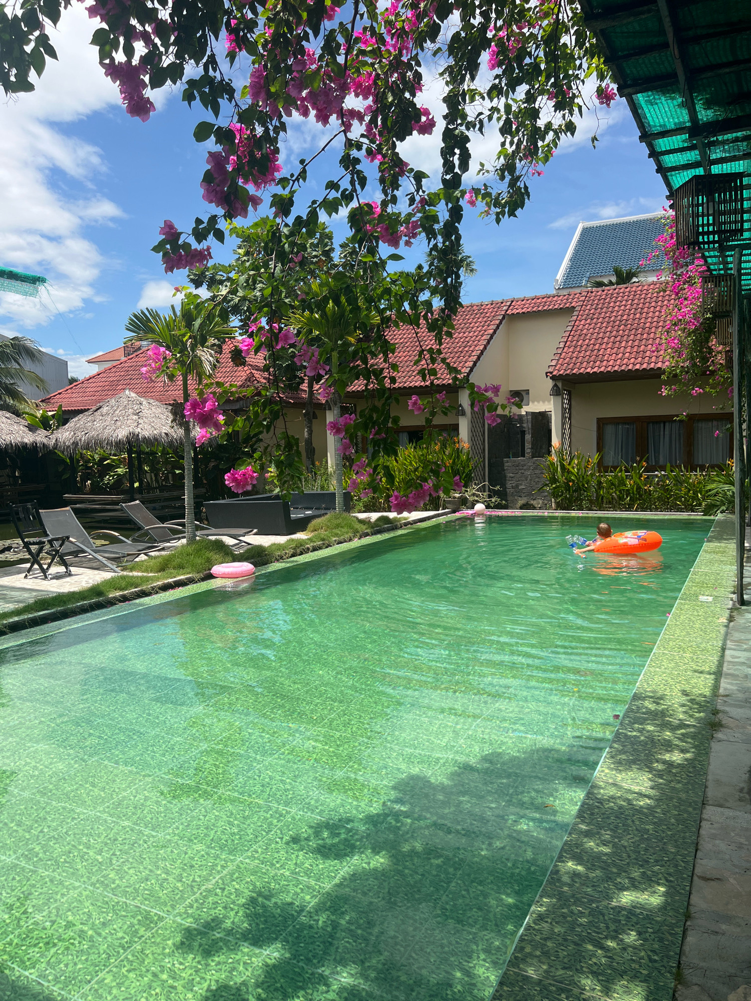 The image depicts a serene outdoor swimming pool area, likely in Hoi An, Vietnam, characterized by lush vegetation and tropical ambiance. In the foreground, a vibrant green swimming pool dominates the scene, its tiled bottom creating a textured mosaic effect. A child, wearing an orange inflatable ring, is visible in the water, seemingly enjoying a swim. Nearby, a pink inflatable ring floats on the water's surface.
The poolside is lined with manicured grass and stone pathways. Several lounge chairs are arranged on the grass, inviting relaxation. In the background, traditional thatched-roof huts, indicative of Southeast Asian architecture, stand alongside modern residential buildings with red-tiled roofs. Pink bougainvillea flowers cascade from overhanging branches, adding a splash of color to the scene. The sky above is a brilliant blue, dotted with fluffy white clouds, suggesting a clear, sunny day. The overall impression is one of a tranquil resort or private residence, offering a peaceful escape from the city bustle.
