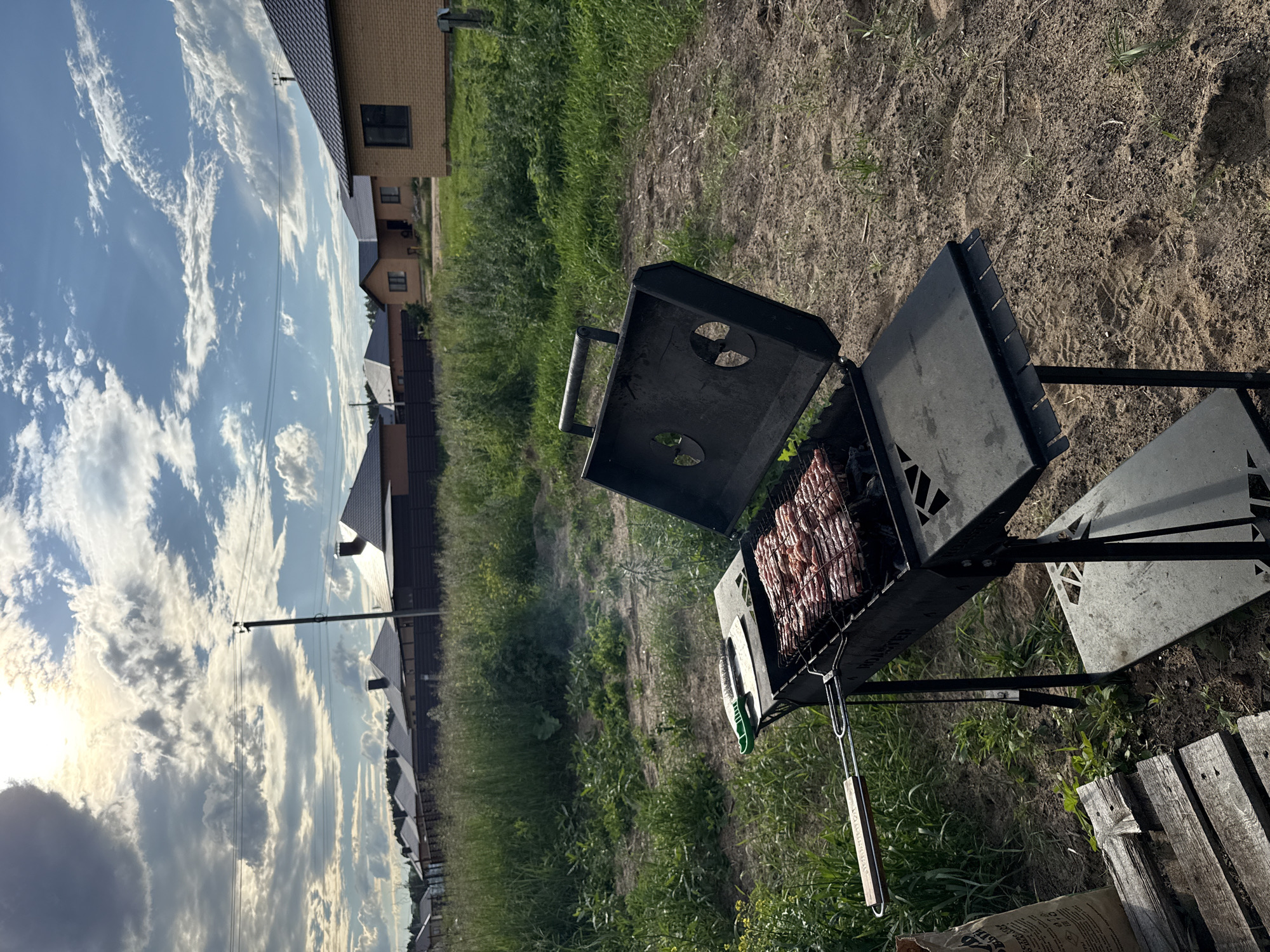 The image captures an outdoor barbecue scene in Chaikovsky, Russia. The primary subject is a black metal grill, open and actively cooking what appears to be rows of meat on a grate. Smoke rises from the grill, indicating it is in use. Next to the grill, a wooden-handled barbecue tool is visible, resting on the side.
The setting is a grassy yard with scattered patches of dirt. In the background, several brick houses with tiled roofs are visible, suggesting a residential area, possibly a dacha or a rural settlement. The sky is a prominent feature, with dramatic, wispy white clouds set against a bright blue expanse. Sunlight is filtering through the clouds, casting a warm glow and creating a sense of late afternoon or early evening.
The overall mood is relaxed and domestic, indicative of a leisurely outdoor meal or gathering. The presence of houses and the well-maintained grill suggest a typical summer activity. No people are directly visible in the frame, but the scene implies their presence, preparing or enjoying food. The image is oriented vertically, with the sky occupying the left portion and the grill and houses on the right. There is some visible text on a brown bag at the bottom of the image, but it is largely illegible.