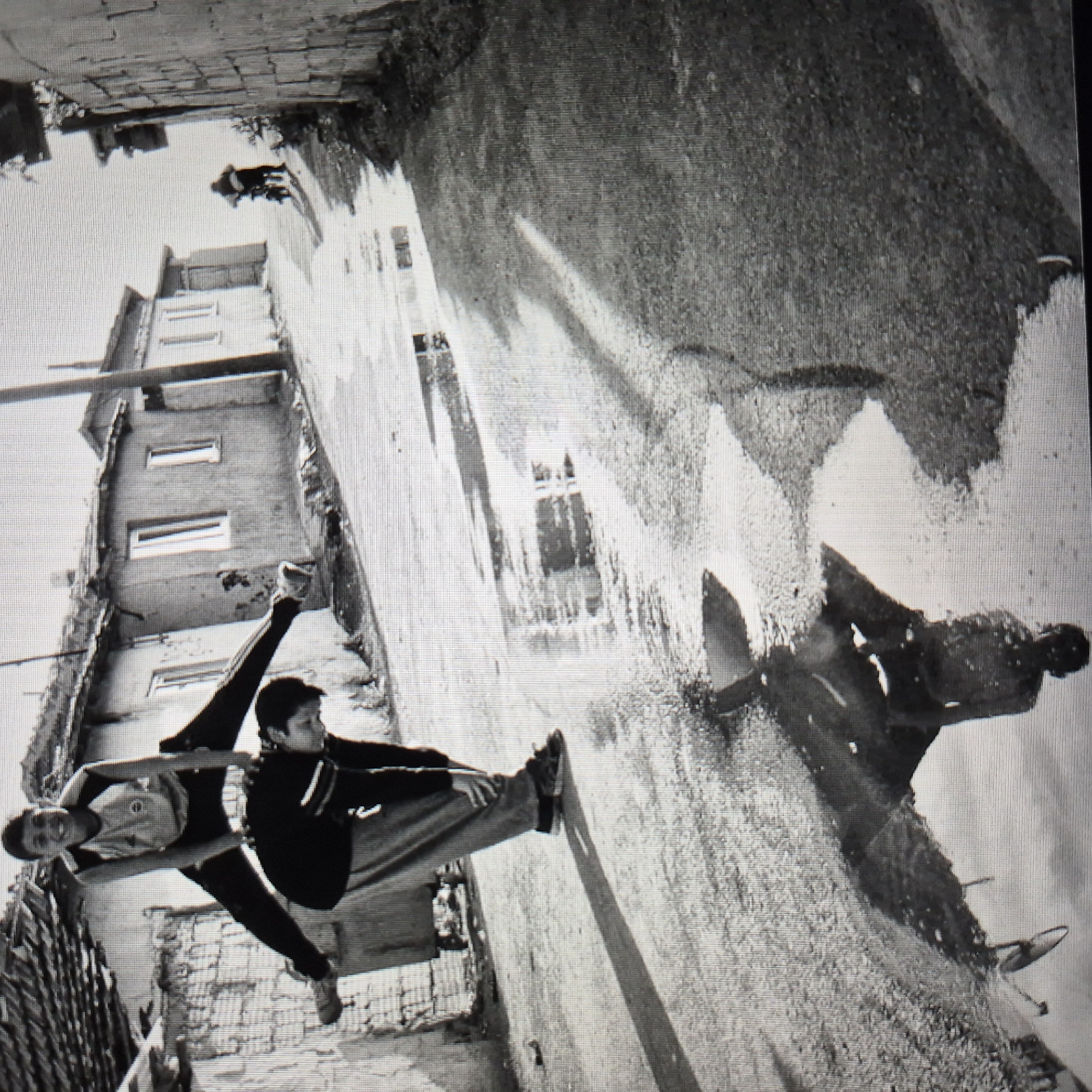 In a black and white photograph, two young boys are engaged in a game of leapfrog on a wet, paved street in Başakşehir, Turkey. The boy on top, with his legs spread wide, is in mid-air, smiling and looking at the camera. The boy below, crouched down, is supporting him and also looking down. The street is lined with modestly built houses, some with damaged walls and tiled roofs, suggesting a less affluent area. Puddles of water on the street reflect the surroundings, including the boys and the houses, adding a sense of depth and atmosphere to the scene. In the background, a person is seen riding a motorcycle, further indicating the urban setting. The overall impression is one of childhood play and resilience amidst a humble urban environment.