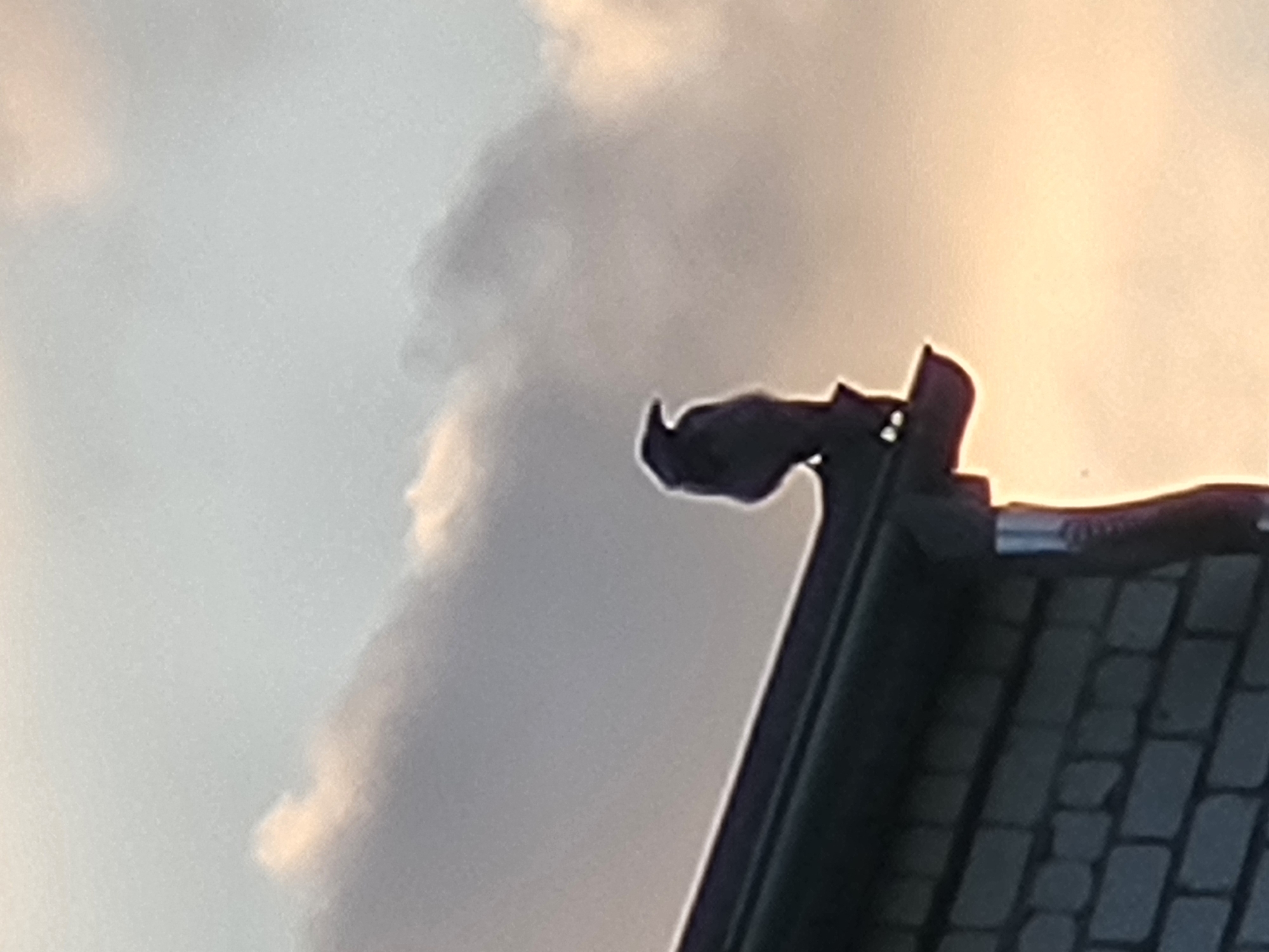 A silhouette of a raven is perched on the edge of a dark-colored gutter system, which runs along the top of a brick building. The building's brickwork is visible in the lower left quadrant of the image, with dark mortar lines between the lighter bricks. The sky behind the bird and building is filled with diffused, soft clouds, suggesting either sunrise or sunset, with a warm, pale orange glow emanating from the right side of the frame. The overall lighting is subdued, creating a dramatic silhouette effect for the bird and the roofline. The location is Ozolnieki, Latvia.