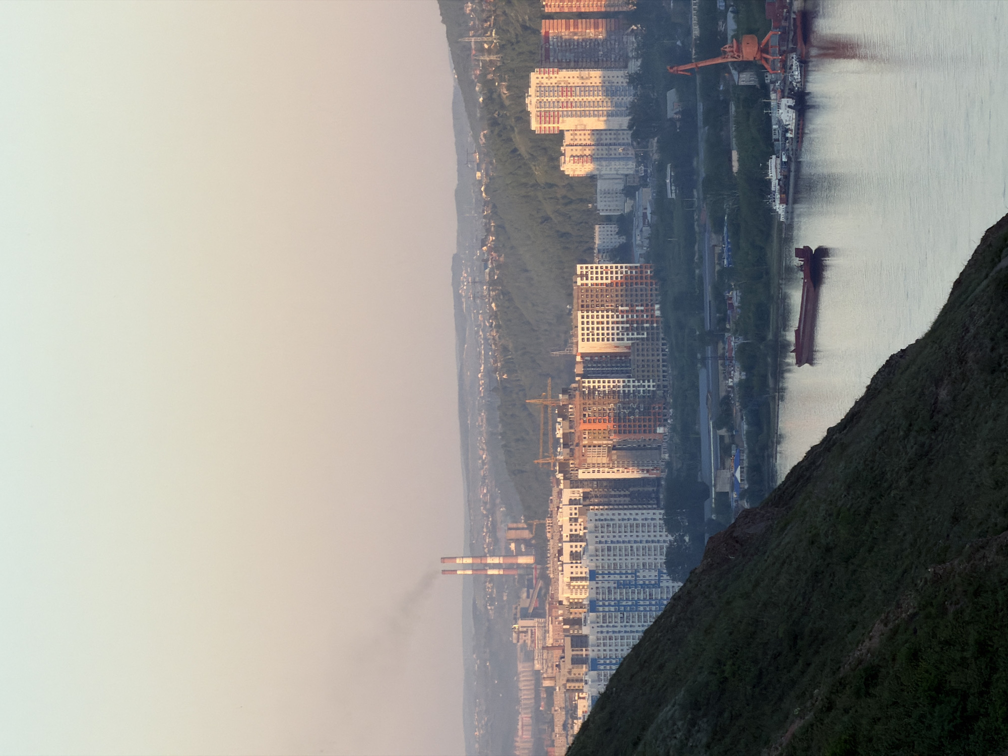 This image presents a panoramic view of the city of Krasnoyarsk, Russia, specifically looking west across the Yenisei River from an elevated position on the right (east) bank, likely from a vantage point such as Karaulnaya Gora or a similar hill.
In the foreground, a steep, dark green, grassy hillside with exposed patches of earth and rock occupies the lower left quadrant. Below this slope, the wide, calm surface of the Yenisei River flows through the midground, reflecting the pale, warm colors of the sky. A long, dark red barge floats stationary or slowly on the river, positioned towards the center-left. Further along the river's right bank, a small port or dock area is visible, with several smaller boats, primarily white and grey, moored alongside the shore. A large, prominent orange gantry crane stands tall on the riverbank at the far right.
Across the river, on the left bank of Krasnoyarsk, a dense urban landscape unfolds. Numerous high-rise residential buildings are visible, some featuring blue-and-white exteriors, while others display warmer orange and brown tones, with several appearing to be under construction or newly completed. In the central background, two tall, slender industrial chimneys, distinctively striped in red and white, emit a stream of dark smoke that drifts to the left. These chimneys are characteristic of the Krasnoyarsk Thermal Power Plant-1 (TPP-1). Adjacent to the power plant, other industrial structures are visible. Beyond the immediate urban development, the landscape transitions into undulating, forested hills, sparsely dotted with additional residential buildings that recede into the distant haze.
The sky above is clear, displaying a subtle gradient from a pale blue-green at the top to a soft peach-pink hue near the horizon, indicative of either sunrise or sunset. The entire scene is bathed in a soft, warm light, illuminating the river and casting a gentle glow on the visible facades of buildings. No individual people or explicit human actions are discernible.