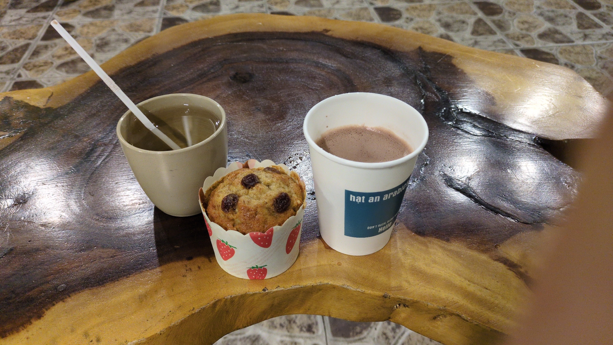 On a varnished wooden table with visible grain, a ceramic cup of clear liquid with a white straw sits next to a strawberry-patterned paper cup containing a raisin muffin. A white paper cup filled with a brown beverage is placed to the right of the muffin. The location is Da Lat, Vietnam, based on the "Hat an arabica" text on the paper cup. The table is likely in a cafe or similar establishment.