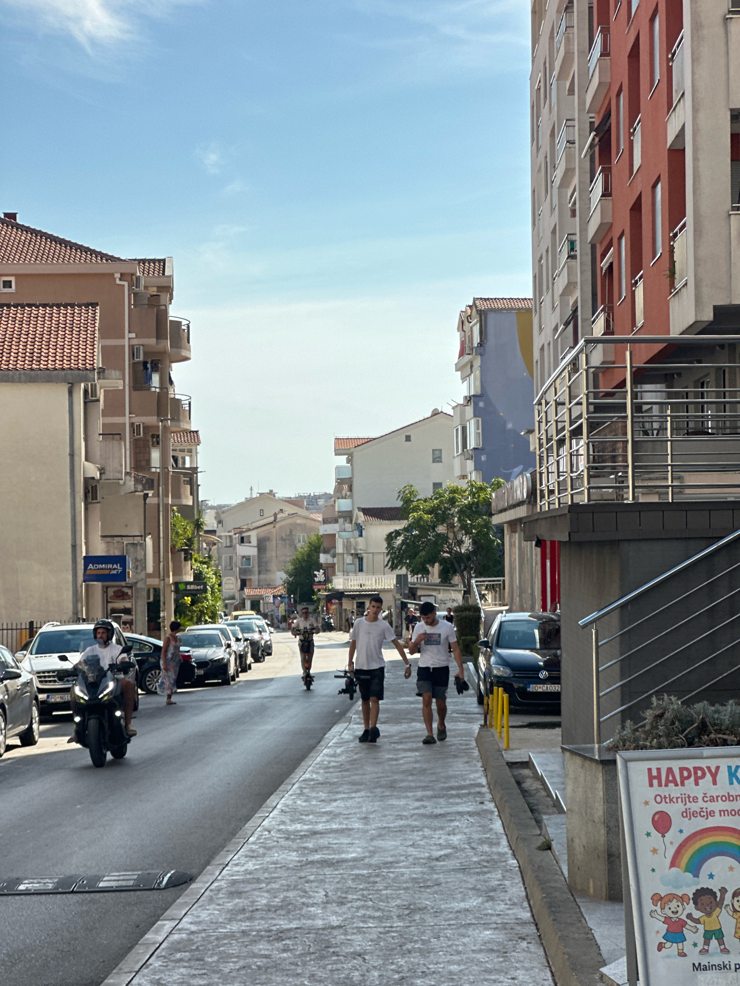 In Budva, Montenegro, on a sun-drenched street, a busy scene unfolds. A man rides a black scooter down the middle of the road, navigating past parked cars. Two young men walk hand-in-hand on the sidewalk, their casual attire suggesting a leisurely day. Further down the street, two other individuals ride electric scooters, adding to the lively atmosphere. Buildings line both sides of the road, their facades a mix of modern apartment blocks and older structures, some adorned with balconies. A banner on the right side of the image promotes "HAPPY K" with colorful illustrations of children and a rainbow, hinting at nearby activities or services. The sky above is a clear blue, with wispy clouds adding a touch of detail to the serene backdrop.