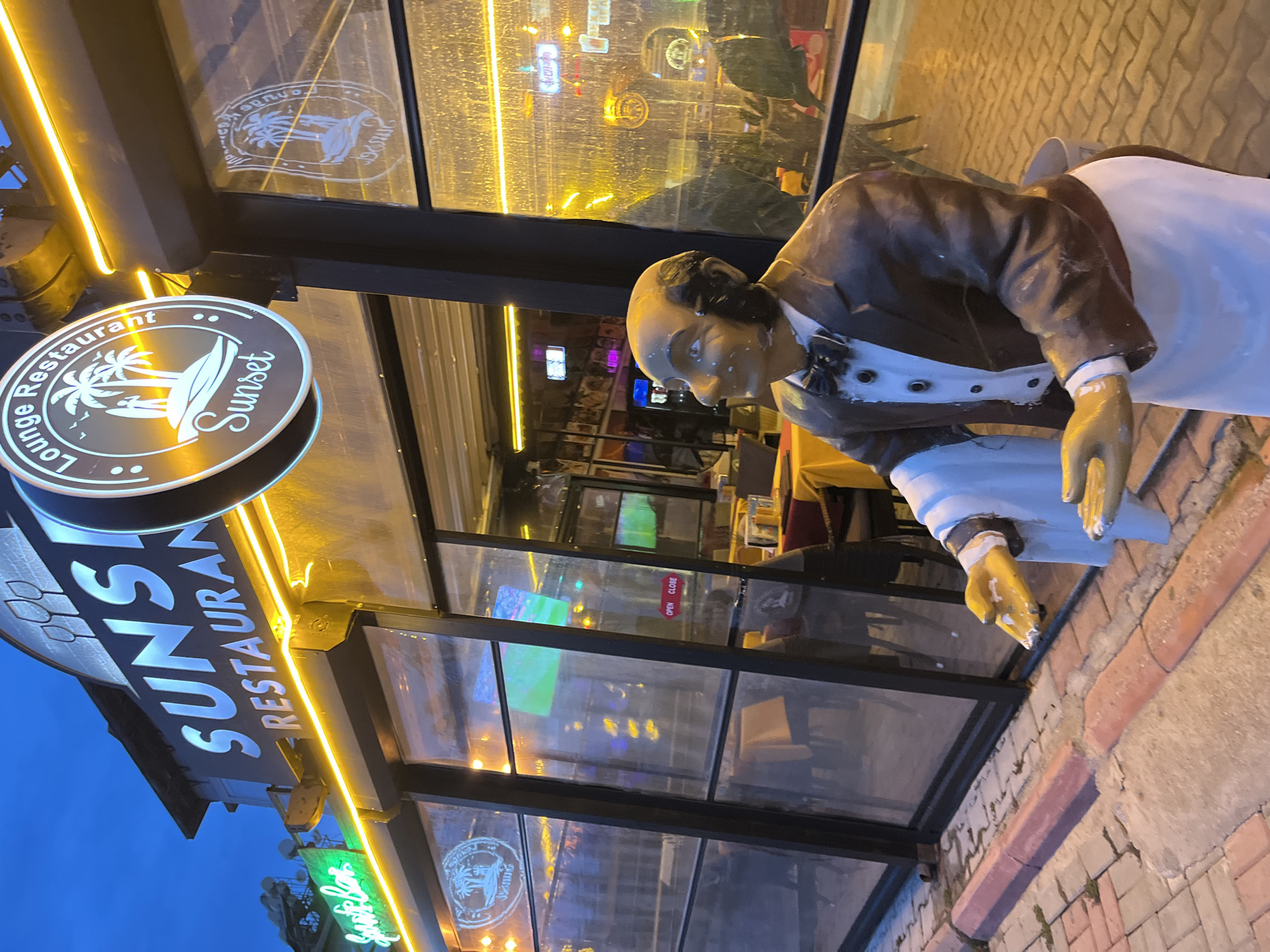 A statue of a waiter stands outside a restaurant. The restaurant, "Sunset," is located in Alanya, Turkey. It is dusk. The restaurant has a large, modern facade with glass walls and a black frame. Neon lights illuminate the restaurant sign. The statue is dressed in a brown suit and white apron, holding a tray. The statue appears worn. The sidewalk in front of the restaurant is made of brick. Inside the restaurant, tables and chairs can be seen. A television is visible, displaying a sports game.