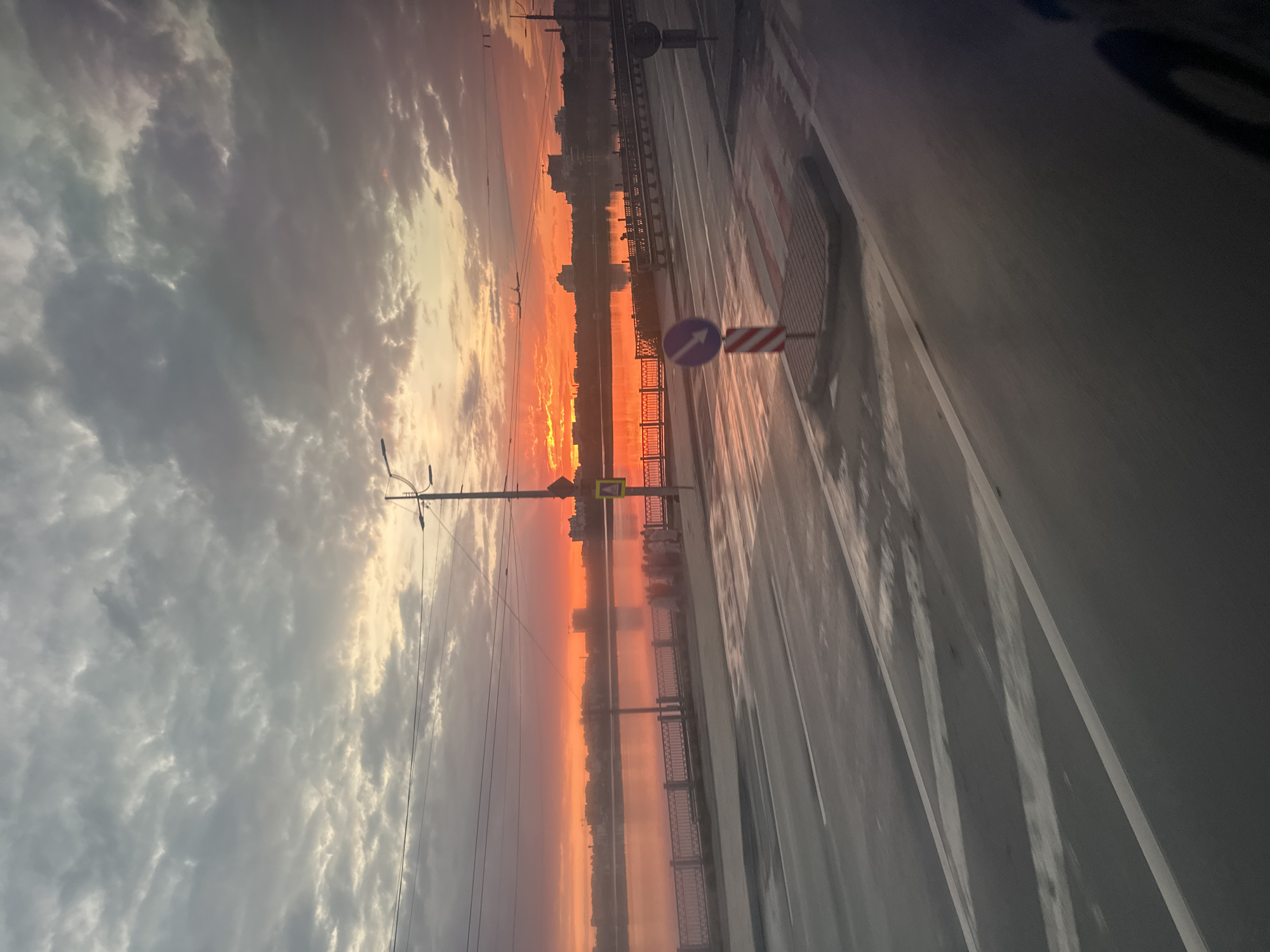This is an outdoor, eye-level, medium shot photograph taken at sunset. The sun is low on the horizon, casting a vibrant orange and pink glow across the sky and reflecting on the water of a river or large body of water. Silhouetted against the bright sky are buildings of a city skyline. In the foreground, a wet asphalt road with white lane markings is visible. There is a sidewalk with a metal railing bordering the water. A pedestrian crossing is marked on the road. Various traffic signs are present, including a blue circular sign with a white arrow pointing left, and a yellow diamond-shaped sign. A lamppost stands on the right side of the image. The clouds are heavy and dark grey, creating a dramatic contrast with the sunset colors. The overall scene suggests a moment of transition from day to night in an urban environment, likely along a waterfront promenade or bridge in Ternopil, Ukraine.