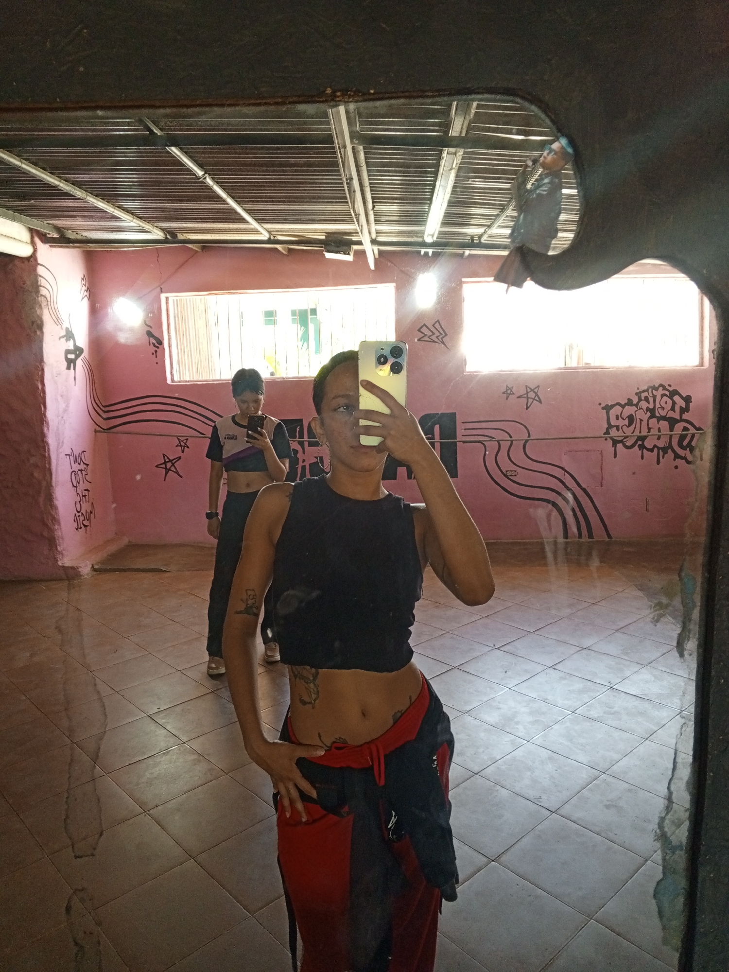 A person is taking a selfie in front of a mirror. The person is wearing a black top and red and black pants. The person's face is partially obscured by their phone. In the background, another person is standing and holding a phone. The room has pink walls with black graffiti. There are two windows in the background. The ceiling has metal beams. The location is a building in Las Barrancas, Venezuela. The floor is tiled.