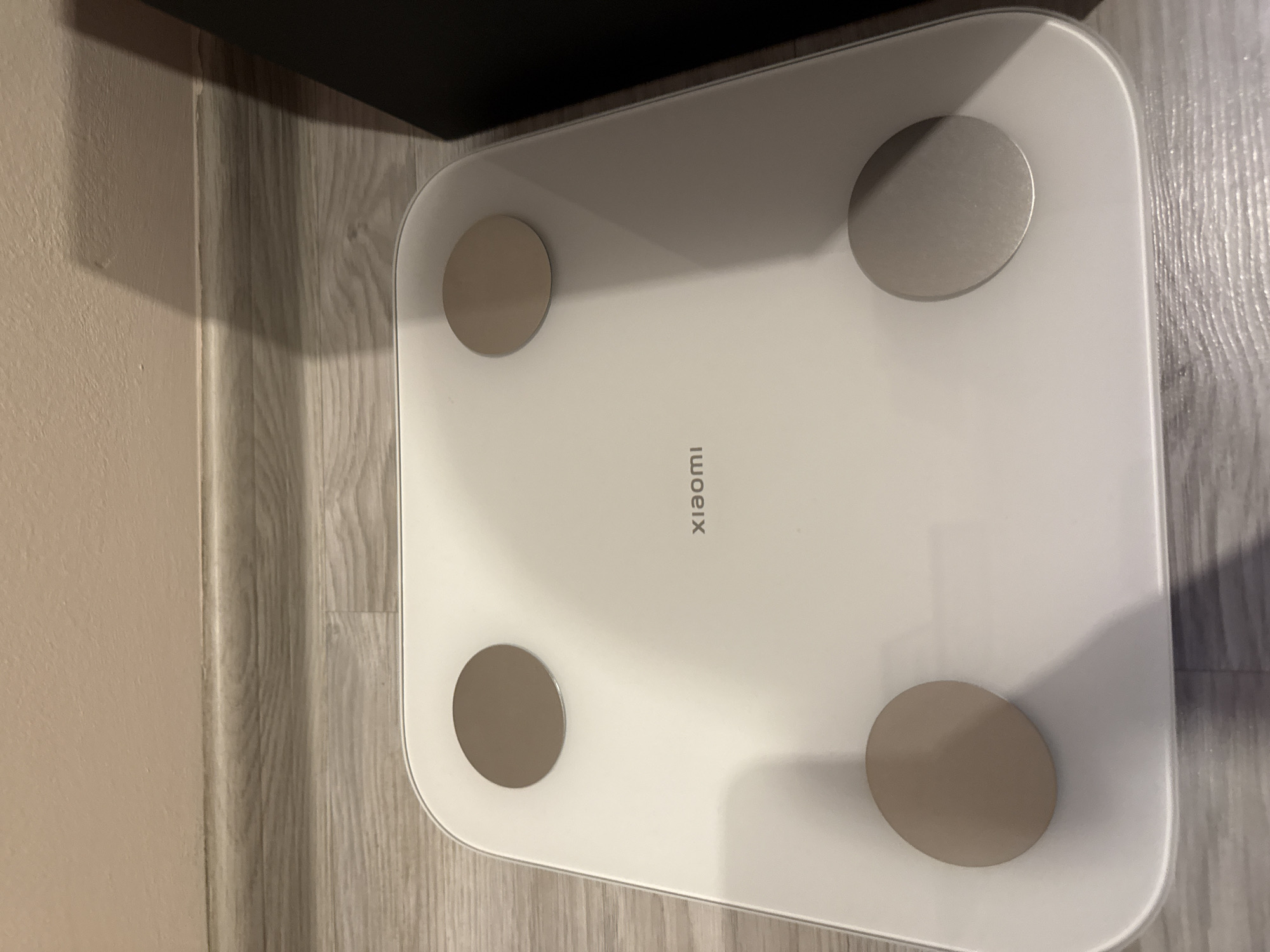 The image shows a white Xiaomi Mi Body Composition Scale 2. It is placed on a wooden-patterned surface next to a light beige wall and a dark object. The location is not discernible from the image.
