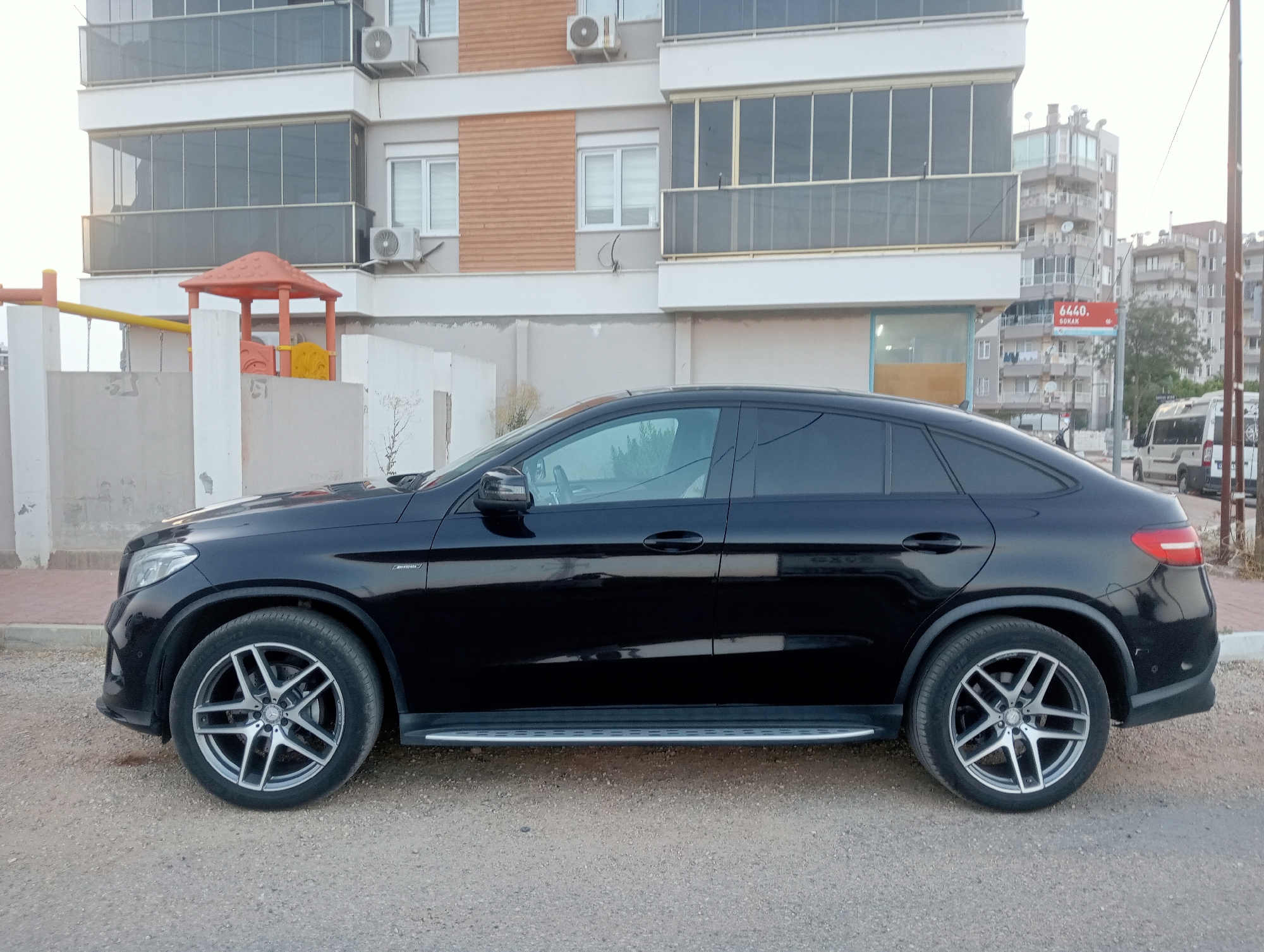 A black Mercedes-Benz coupe SUV is parked on a gravel lot next to a sidewalk. The vehicle is oriented to the right, with its side facing the camera. The car has a sunroof, tinted windows, and chrome accents. In the background, there is a multi-story building with balconies and air conditioning units. A small playground with an orange slide and yellow swing set is visible next to the building. Further in the background, other buildings and a street sign are visible. The sky is overcast.