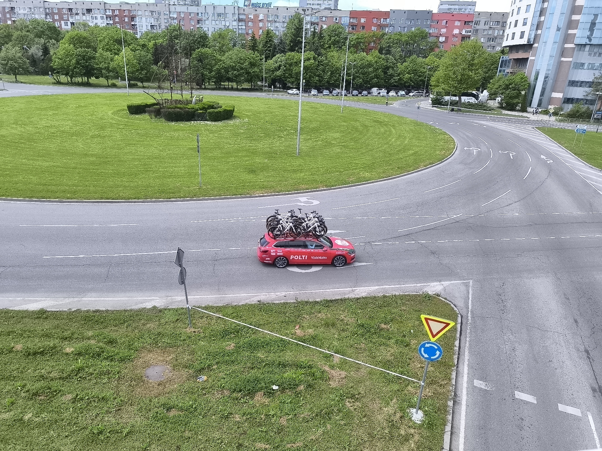 The image shows a view from above of a road intersection in Burgas, Bulgaria. A red car with multiple bicycles strapped to its roof, labeled "POLTI VeloMaIra," is positioned near the lower center of the frame. A roundabout is to the upper left of the car. The road leading from the bottom right of the frame has a "yield" sign and a "mandatory roundabout" sign. In the background, there are buildings, trees, and parked cars visible. A sculpture is visible within the roundabout. The weather appears to be overcast.