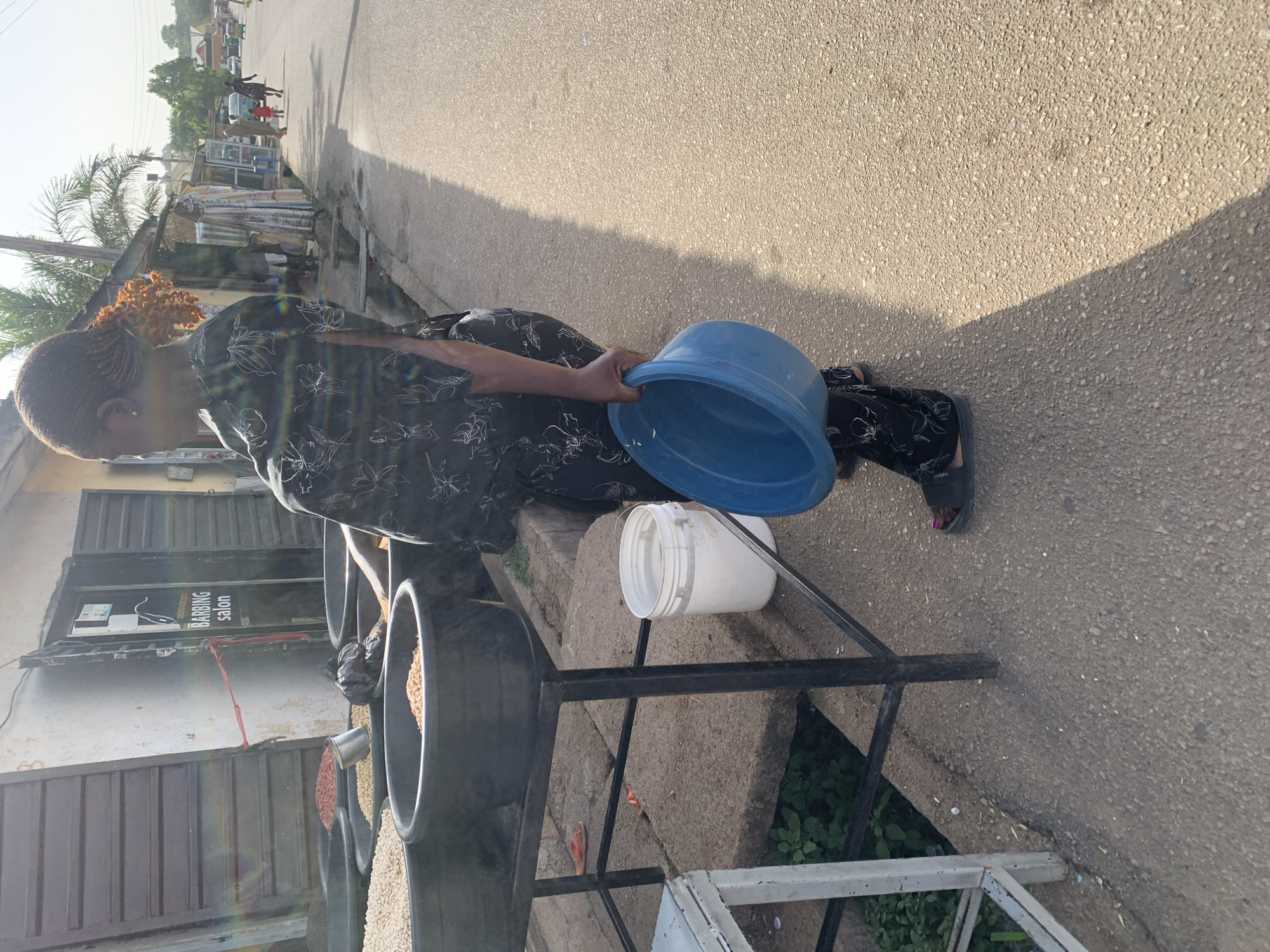 A woman in Gura Top, Nigeria is holding a blue plastic bowl over a white plastic bucket, which sits on a black metal frame. Several large, black circular containers filled with what appears to be grains or beans are also on the frame. The "Barbing Salon" shop is visible in the background. The scene is outdoors on what appears to be a paved road or sidewalk.