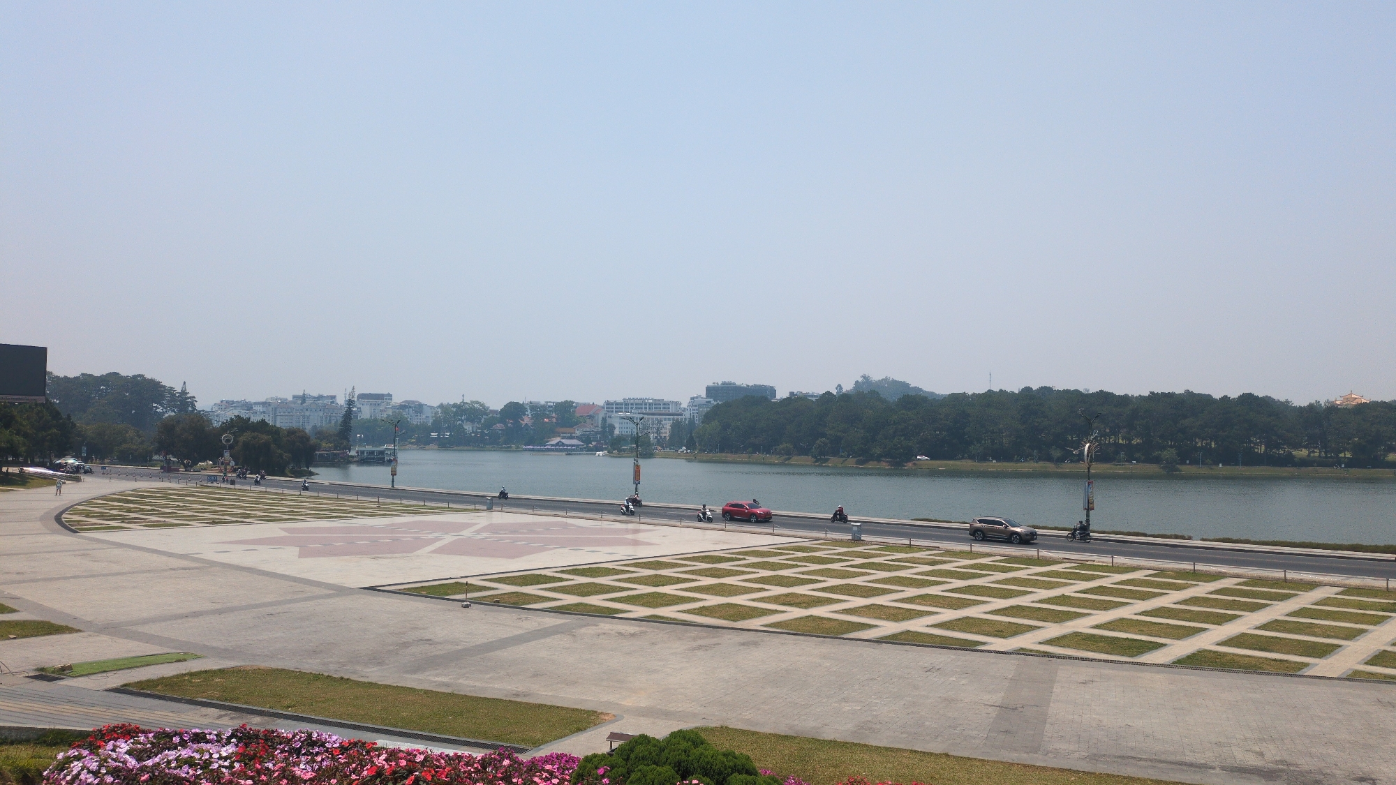 The image shows a wide-angle view of a plaza overlooking a lake in Da Lat, Vietnam. In the foreground, the plaza is composed of concrete and interspersed with square patches of grass and flowerbeds. A pattern of painted arrows is visible in the plaza. A roadway runs along the edge of the lake, with a red SUV and a brown SUV traveling along it, along with several motorcycles. The lake is bordered by trees and buildings in the background. The sky is overcast. Several light posts are visible along the roadway.