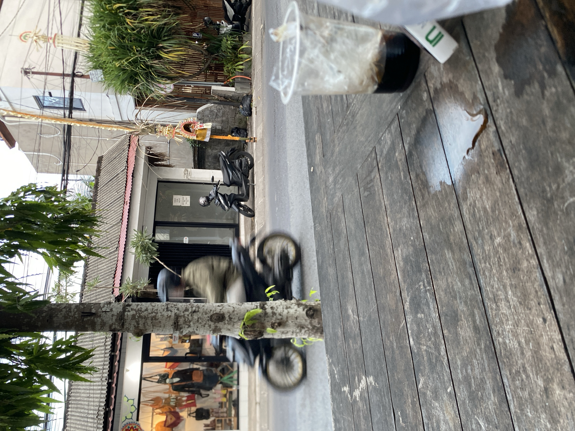 This media file depicts a scene on a street in Mengwi, Indonesia. The orientation of the image is landscape. The foreground features a weathered wooden table with spilled liquid, likely coffee or tea, forming a dark puddle. A clear plastic cup with a straw and ice is visible on the table, suggesting a beverage was recently consumed.
Beyond the table, a street runs parallel to the viewer's perspective. Several objects are present in the middle ground. A black motorcycle is parked on the side of the road. Behind the motorcycle, a building with a gray corrugated roof is visible, with a storefront featuring bags and other merchandise displayed inside. Another building with a white facade and a small, dark-tinted window is situated to the left of the corrugated-roof building. Traditional Balinese decorations, possibly for a festival or ceremony, are displayed on tall wooden poles along the roadside. These decorations are elaborate and crafted from natural materials. A lush green plant is situated in front of the buildings.
In the background, trees and more buildings can be seen. The image has a slightly blurred effect, particularly the moving elements like a person on a scooter. This suggests the photo may have been taken during the daytime. The overall atmosphere appears casual and everyday, with the presence of a street, shops, and a beverage on a table. The weather appears to be clear, as indicated by the lighting. No specific emotions are conveyed, but the scene suggests a typical street view in a tropical urban environment. There is some visible text on a white label attached to the plastic cup, but it is illegible. There are also small, illegible signs on the shop windows.