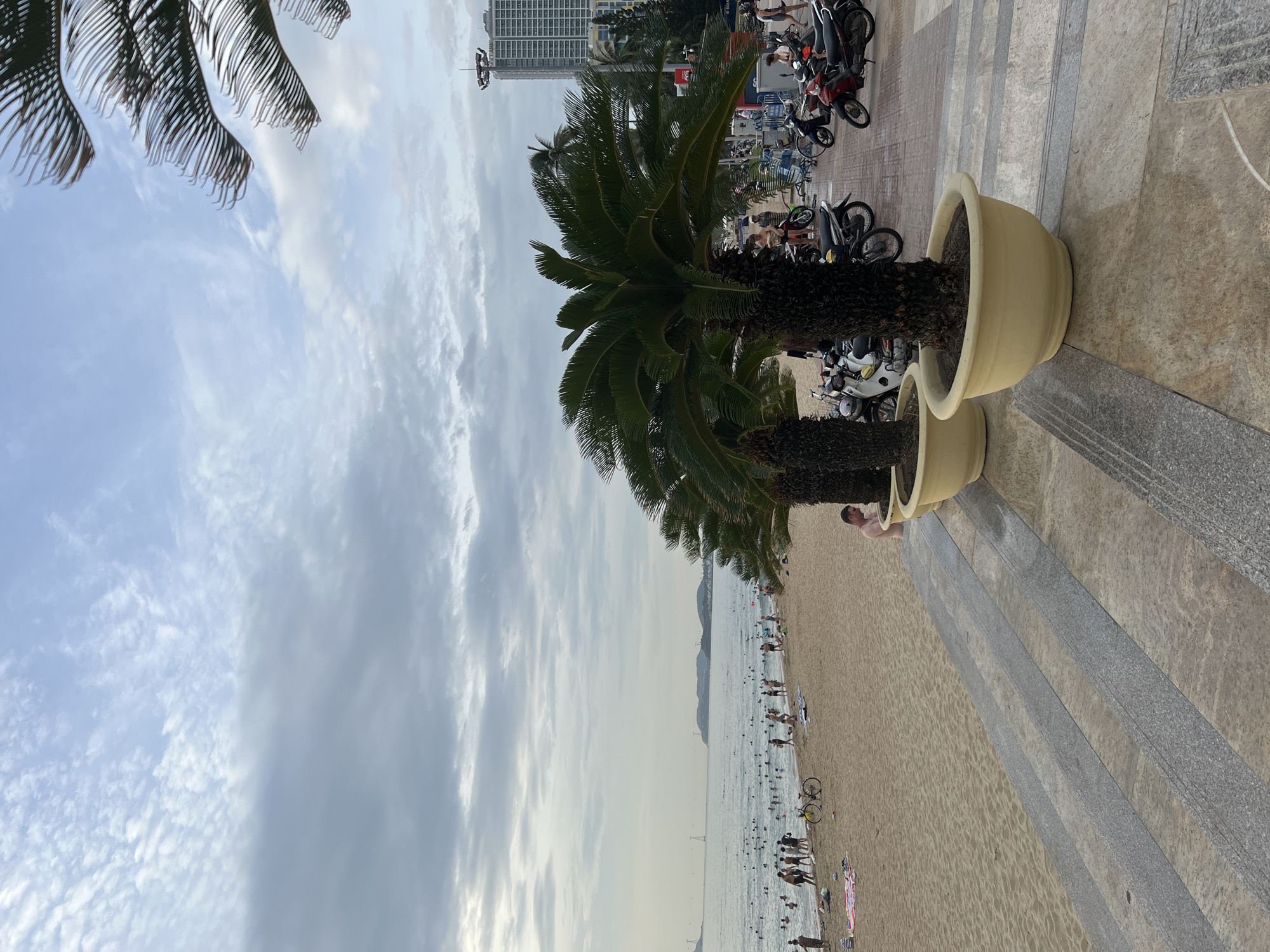 The image depicts a crowded beach scene in Nha Trang, Vietnam, likely along Tran Phu Beach, a popular stretch known for its sandy shores and promenade. The sky is overcast with scattered clouds, suggesting a diffused light condition. In the foreground, large potted palm trees line a raised walkway paved with stone tiles, some of which are wet. Beyond the walkway, a wide sandy beach is teeming with people. Many individuals are in the water, swimming or wading, while others relax on the sand, sunbathe, or stroll along the shore. Beach towels and a bicycle are visible on the sand. Further back, the ocean stretches to the horizon, with distant hills or islands visible. On the right side of the image, a modern high-rise building stands tall, indicating the urban setting of the beach. A collection of motorcycles is parked along the promenade behind the palm trees, a common sight in Vietnamese cities. The overall atmosphere is one of leisure and activity on a busy summer day.