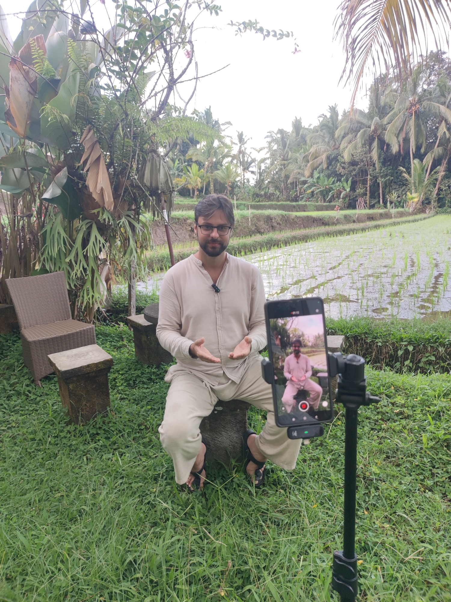 The image depicts a man sitting on a concrete bench in a grassy area near rice paddies. The man is fair-skinned, wearing glasses and a light-colored, long-sleeved shirt and pants, and sandals. He is positioned in front of a smartphone mounted on a black tripod, recording him. The smartphone screen shows a mirrored image of the man in the same pose. Behind him, there's a lush green landscape with various plants and trees, including what appears to be palm trees and other tropical foliage. A rice paddy is visible in the background. To the left, there is a brown woven chair and a small concrete pedestal. The image was taken in Tegallalang, Indonesia.