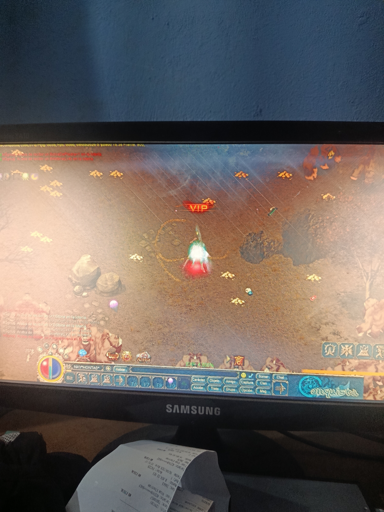 A digital photograph shows a computer monitor displaying the game "Conquista" in Spanish. In the game interface, a character labeled "VIP" is centrally located in an open, sandy area with sparse vegetation and rock formations. Various in-game notifications relating to receiving lottery winnings, are shown. The monitor is a Samsung brand. A partially visible receipt is in the foreground, showing financial information, including a date of April 16, 2026, a transaction for 8,434.53 VES, a card number starting with 422169, and the words "Visa Electron." Juan Griego, Venezuela is not discernible from this image.