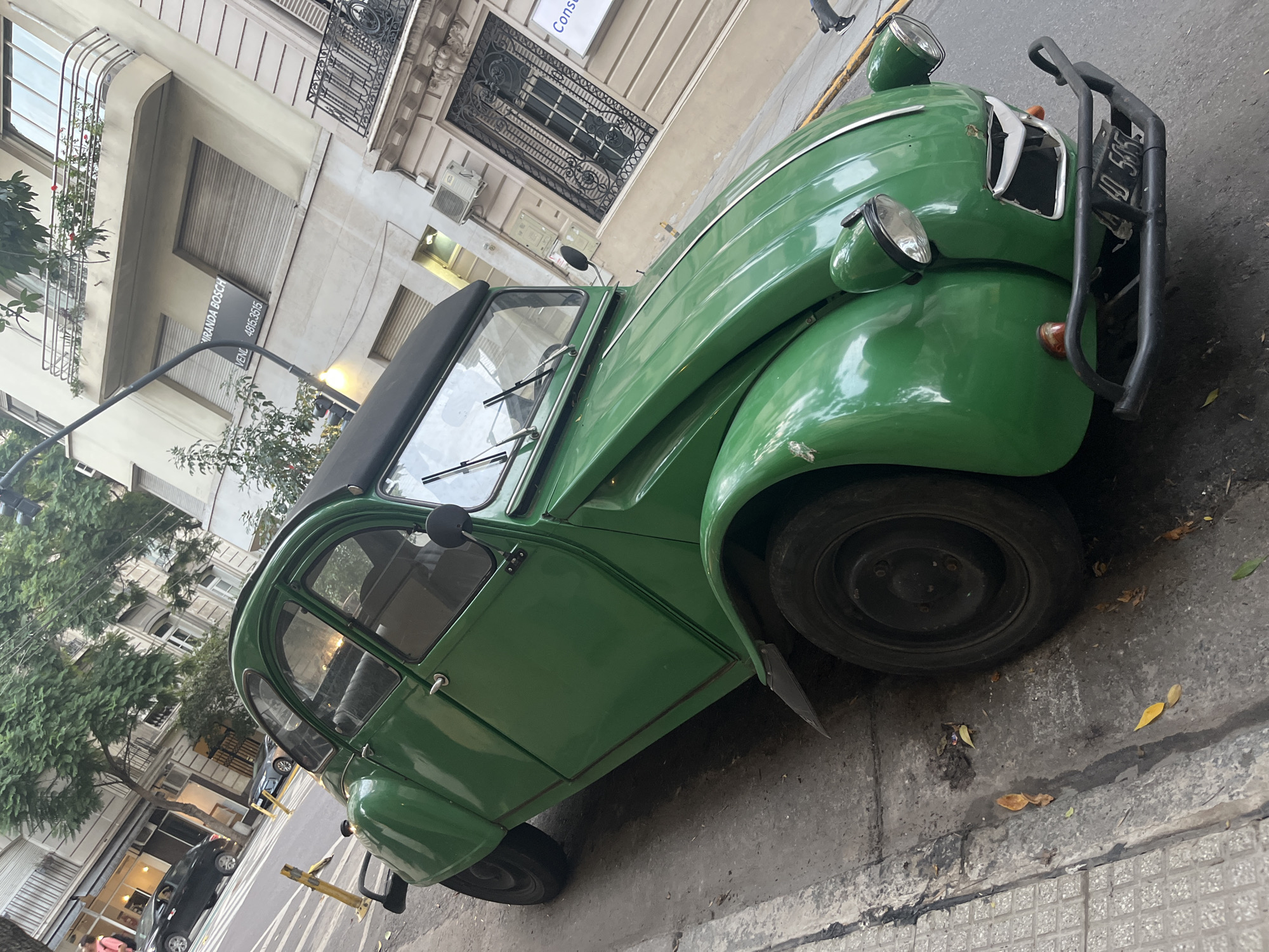 A green Citroen 2CV is parked on a street in Buenos Aires, Argentina. The car is angled towards the right of the frame and occupies a significant portion of the image. The front of the car features a black bumper and a license plate. The car appears to be in a residential area, as buildings with balconies and windows are visible in the background. A street lamp and some parked cars are also noticeable in the background. The sidewalk is visible next to the car. The overall lighting of the scene suggests it is daytime.
