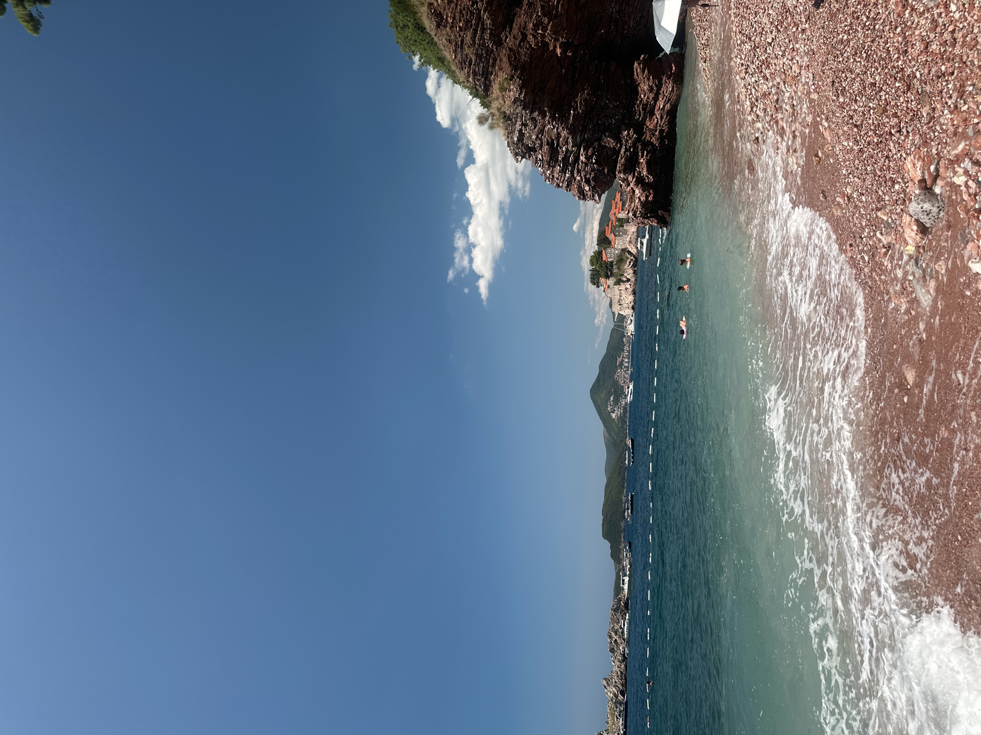 A scenic view of the coast in Sveti Stefan, Montenegro, showcases a clear blue sky and calm turquoise waters. The foreground features a pebble beach with red-tinted stones. In the middle ground, several people are seen swimming or relaxing in the sea, with a white marker line dividing the swimming area from the open water. The background reveals a charming village with red-tiled roofs perched on a rocky island, connected to the mainland by a narrow causeway. Numerous boats are anchored in the bay, and lush green hills rise behind the village. A large, reddish-brown rock formation juts out into the sea on the right side of the frame, partially covered with dry grass. An open umbrella stands on the beach near this rock.