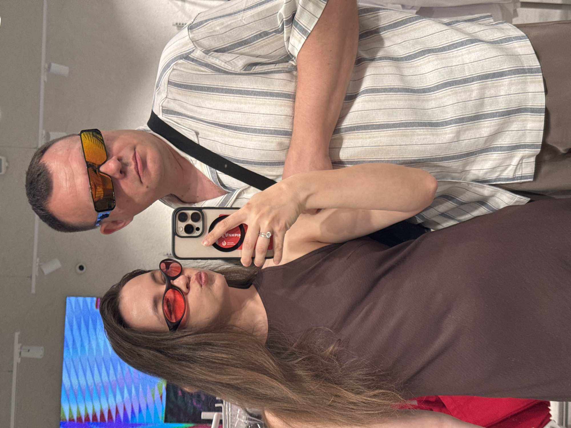 A woman with long brown hair and red-tinted glasses is holding an iPhone 14 Pro, taking a selfie with a man. The man has short hair and is wearing gold-tinted sunglasses. He is wearing a white and blue striped shirt. The woman is wearing a brown dress. In the reflection, a screen with blue and purple graphics is visible in the lower left corner. The photo was taken in a store in the city of Khimki, Russia.