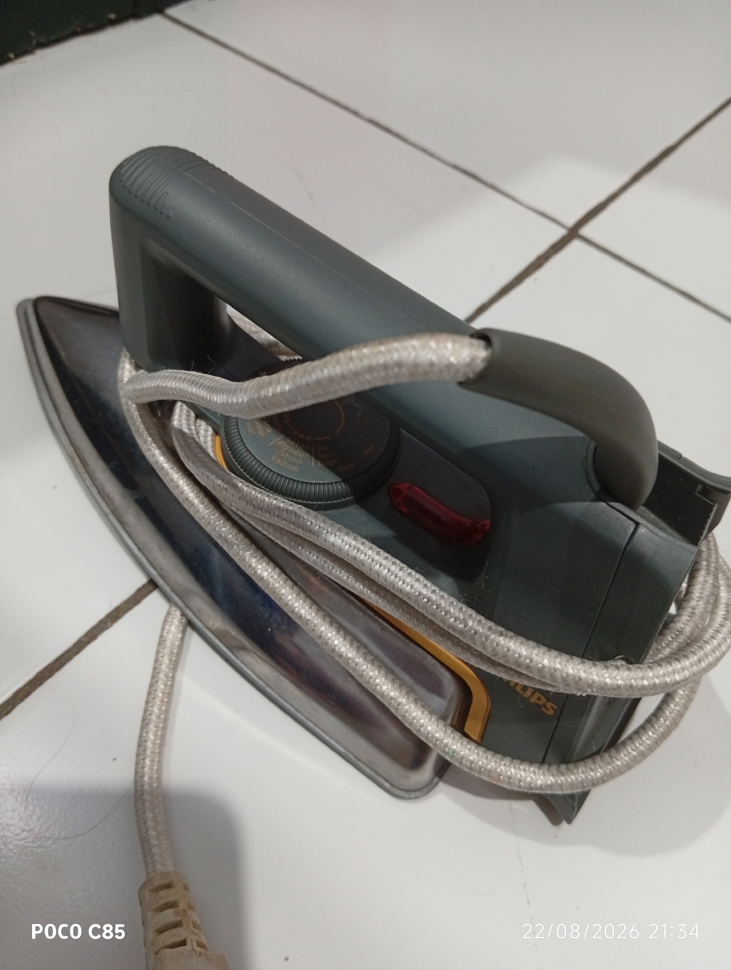 An iron with a dark gray body and silver soleplate rests on a white tiled floor. The power cord, encased in a braided silver material, is coiled around the iron. A red indicator light is visible on the top of the iron, along with a rotary dial for temperature settings. The Philips logo is partially visible on the side of the iron. The tiled floor shows grout lines that form a grid pattern. A date stamp "22/08/2026 21:34" and "POCO C85" are present in the lower right and left corners of the image respectively. The scene is indoors, with the lighting suggesting it is either evening or a dimly lit interior space. This image was taken in South Tangerang, Indonesia.