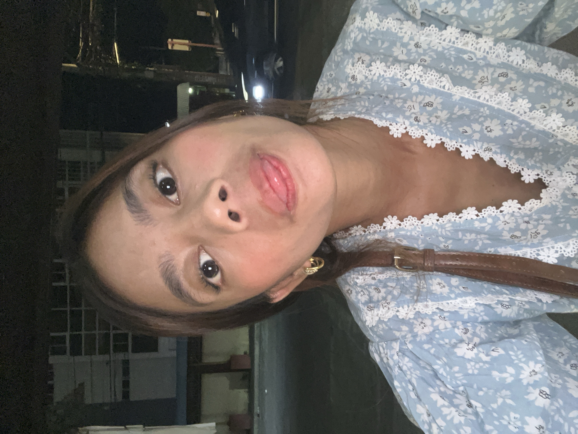 The image is a close-up, portrait-oriented photograph taken outdoors at night, likely in Davao City, Philippines. The subject is a young woman with brown hair, wearing a light blue dress with a white floral pattern and white lace trim. She is looking directly at the camera with her tongue slightly out. Her makeup appears to include blush and mascara. She is wearing a gold hoop earring and a brown belt is visible across her chest. The background is dark, suggesting it is nighttime, and includes some building structures with visible windows and lighting, as well as some trees. A car's headlights are faintly visible in the distance. The overall impression is of a casual, possibly selfie-style photograph taken during an evening outing.