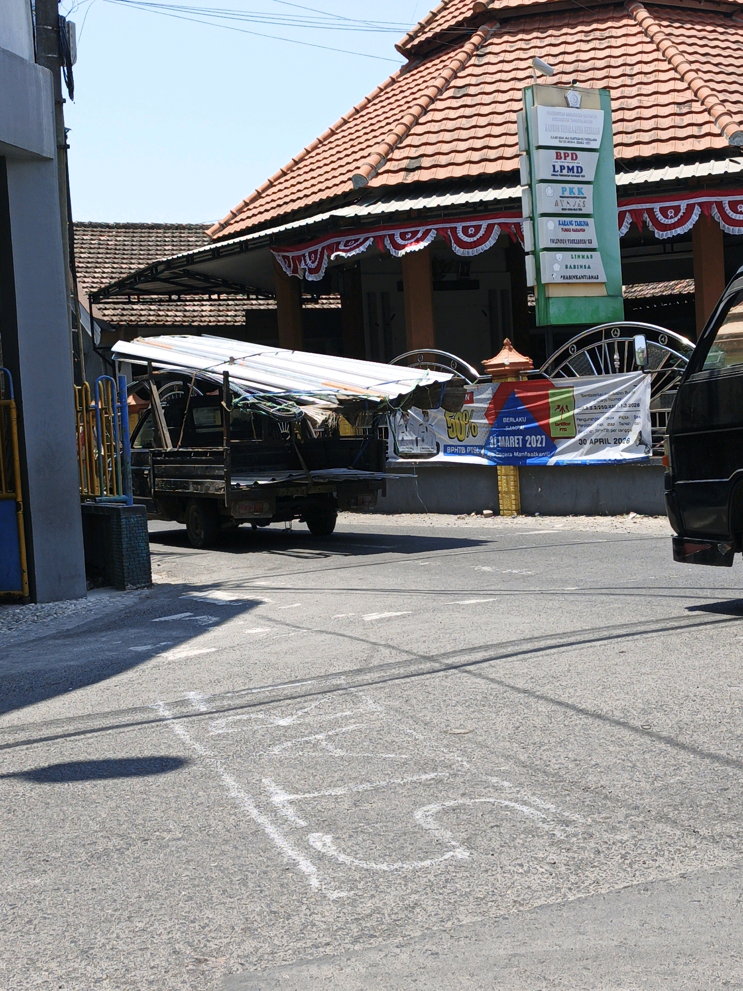 The image shows a street scene in Ketegan, Indonesia. In the foreground, a hopscotch game is drawn with white chalk on the asphalt. A dark-colored flatbed truck, loaded with what appears to be construction materials and a white tarp, is parked on the right side of the street. In the background, a building with a red tile roof and decorative banners is visible. A green directional sign with text in Indonesian is mounted on a pole in front of the building. A black vehicle is partially visible on the far right of the image. The sky is clear and blue.