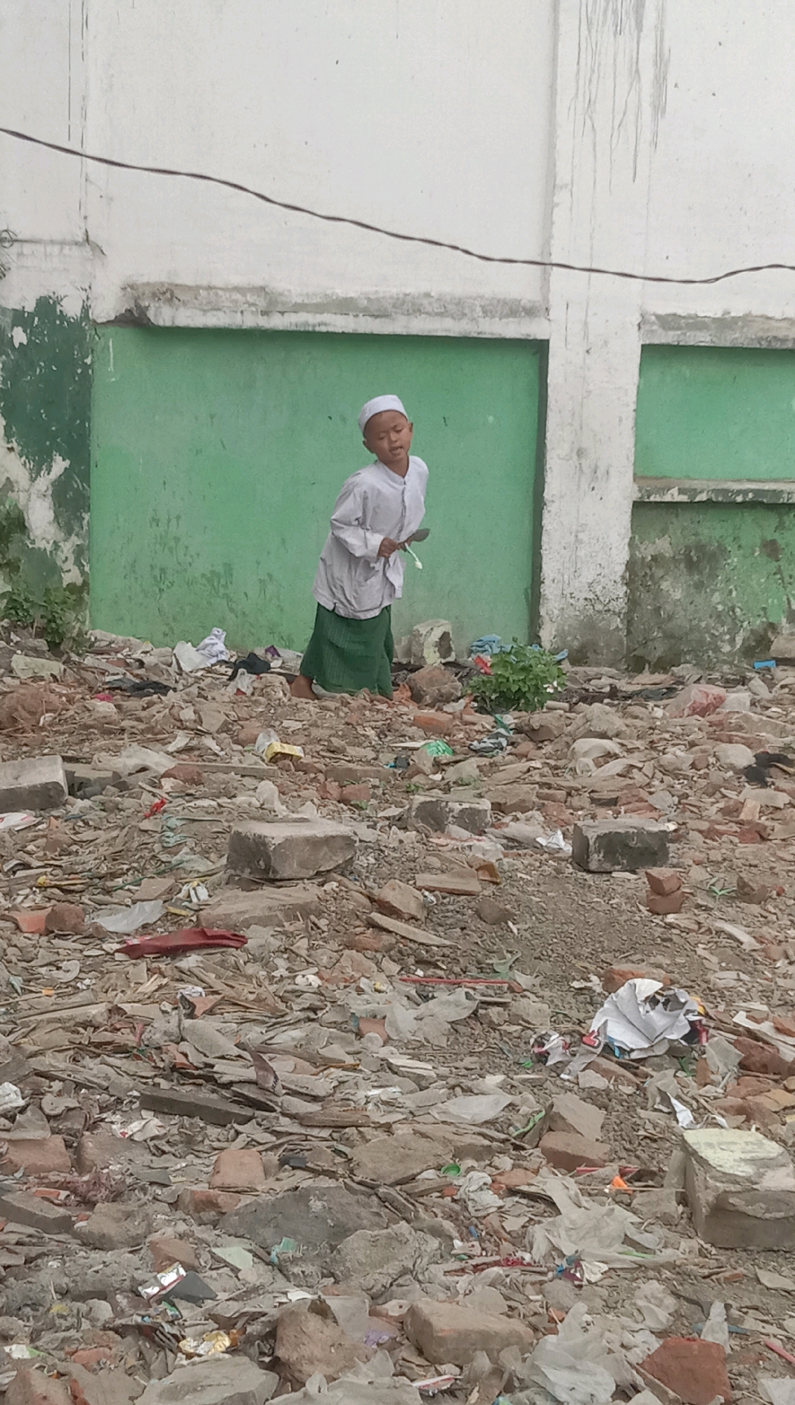 The image depicts a young boy standing in a debris-filled area. The boy is wearing a white skullcap and a white shirt over a green sarong. He appears to be looking towards his right and holding a spoon in his right hand. The ground around him is covered in a mixture of broken bricks, rubble, and scattered debris, suggesting a damaged or demolished structure. In the background, a green wall with white sections and a wire running across it is visible. This scene is located in the city of Geneng Waru, Indonesia.