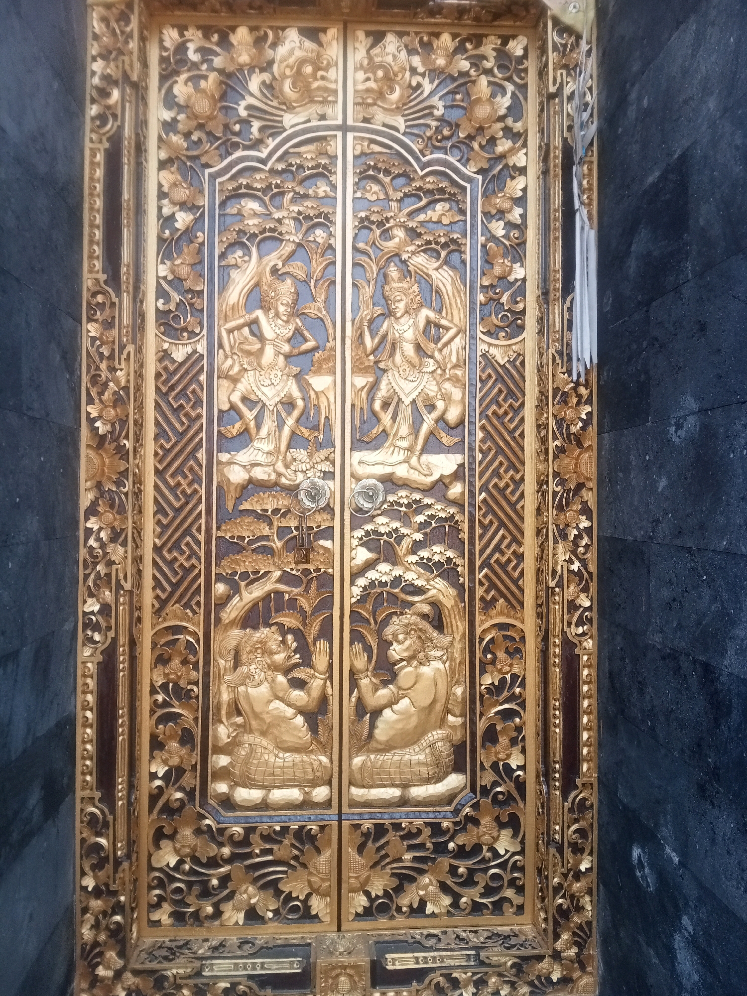 The image shows a closed, ornate wooden door with intricate gold carvings. The door is divided into two panels, each featuring carved figures and decorative motifs. The left panel depicts a seated figure with hands clasped in prayer, surrounded by foliage and trees. The right panel displays a similar seated figure. Above these figures, on each panel, is a carving of a dancer-like figure in mid-movement. The entire door is framed by elaborate golden carvings of floral patterns and geometric designs. The door is set against a dark, textured wall, likely made of stone or dark tile. The overall impression is one of religious or cultural significance, possibly belonging to a temple or a traditional building. The image is a close-up, focusing on the detailed craftsmanship of the door. The location is identified as Mendoyo, Indonesia, suggesting it is likely a cultural artifact from that region.
