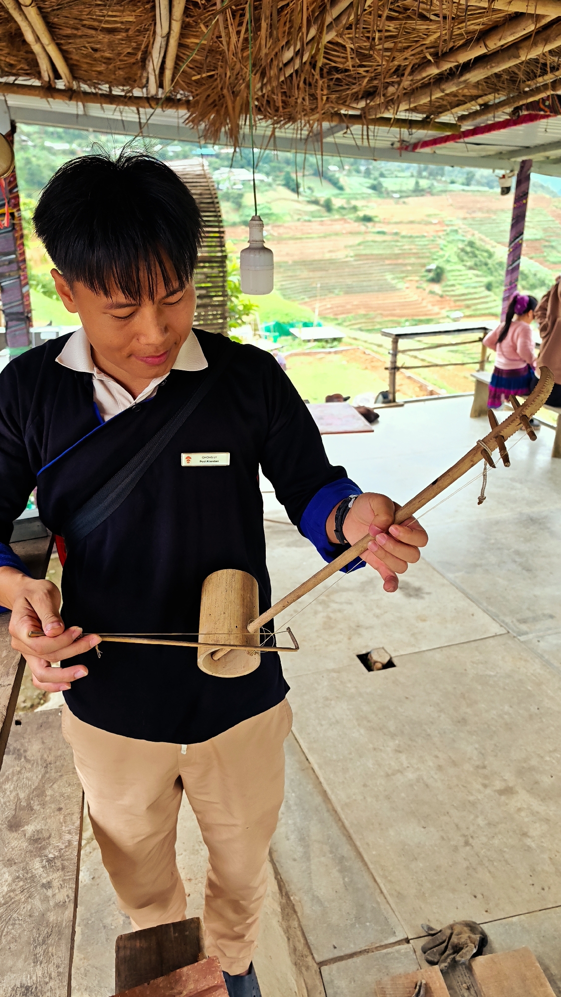An East Asian male is depicted holding and manipulating a bamboo stringed instrument within an open-air, thatched-roof shelter in Dế Xu Phình, Vietnam. The man, appearing in his twenties, has short black hair and wears a black long-sleeved shirt with a white collar and blue cuff accents, paired with light brown trousers. A name tag on his left chest identifies him as "CHONG LY / Poui Khembou". He focuses on the bamboo instrument, which features a cylindrical resonator, a slender neck with tuning pegs, and strings stretched over a rectangular bamboo bridge. The shelter's ceiling is constructed from woven thatch and bamboo, with a white incandescent bulb hanging by a green wire. The concrete floor is visible beneath him. In the background, terraced agricultural fields, characteristic of the mountainous landscape of Dế Xu Phình, stretch across rolling hills. A second female figure in a pink top is partially visible in the distant background to the right.