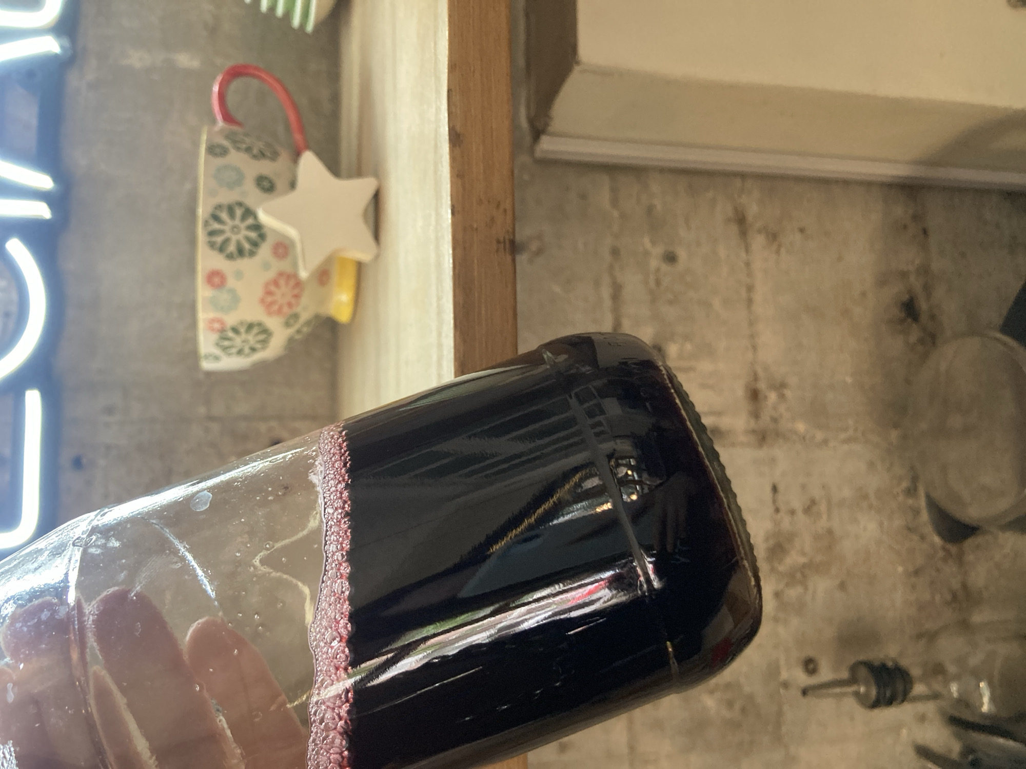 A tilted image shows a glass bottle filled with a dark red liquid, possibly wine, partially obscuring the view of a room. A person's hand is visible through the clear part of the bottle. In the blurred background, there's a wall, a wooden beam, a mug hanging with a star decoration, a neon light, and some kitchen utensils on a counter. The location is an unspecified space within the city of Unknown, Unknown.