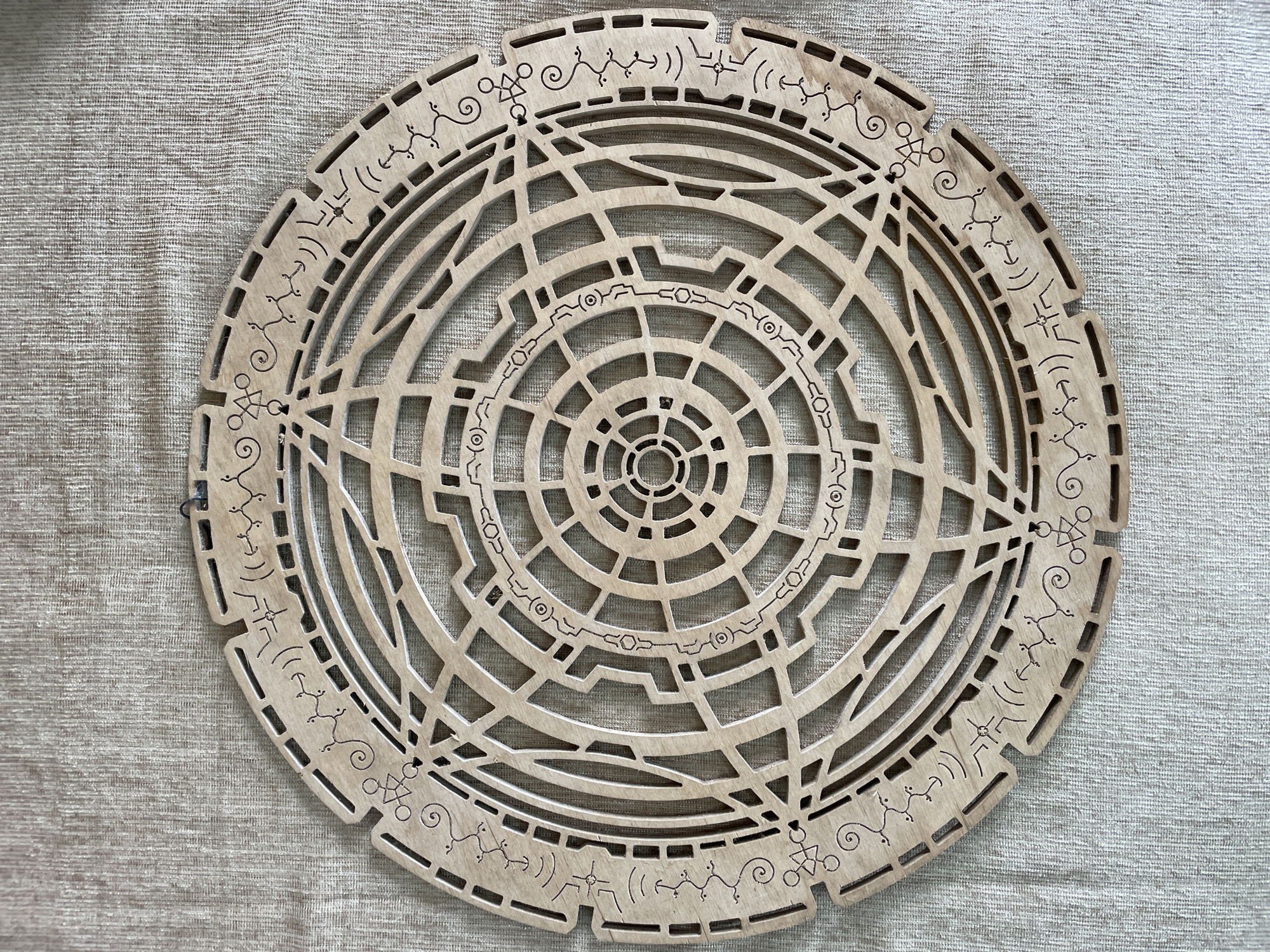The image shows a circular, wooden, laser-cut artifact with intricate geometric patterns and what appear to be symbols or runes. The artifact is placed on a textured, neutral-colored fabric background, possibly a tablecloth or a textured wall covering. The artifact has a layered design with concentric circles and radial lines forming various shapes and openings. The outer edge is notched and adorned with individual symbols and decorative swirls. The colors are monochromatic, primarily light brown wood against the beige background. There are no people or animals present in the image. The lighting suggests an indoor setting, with a slight shadow cast on the fabric, indicating a light source from above or the side. The specific location within the city of Bhagsu, India, cannot be determined from the image alone.