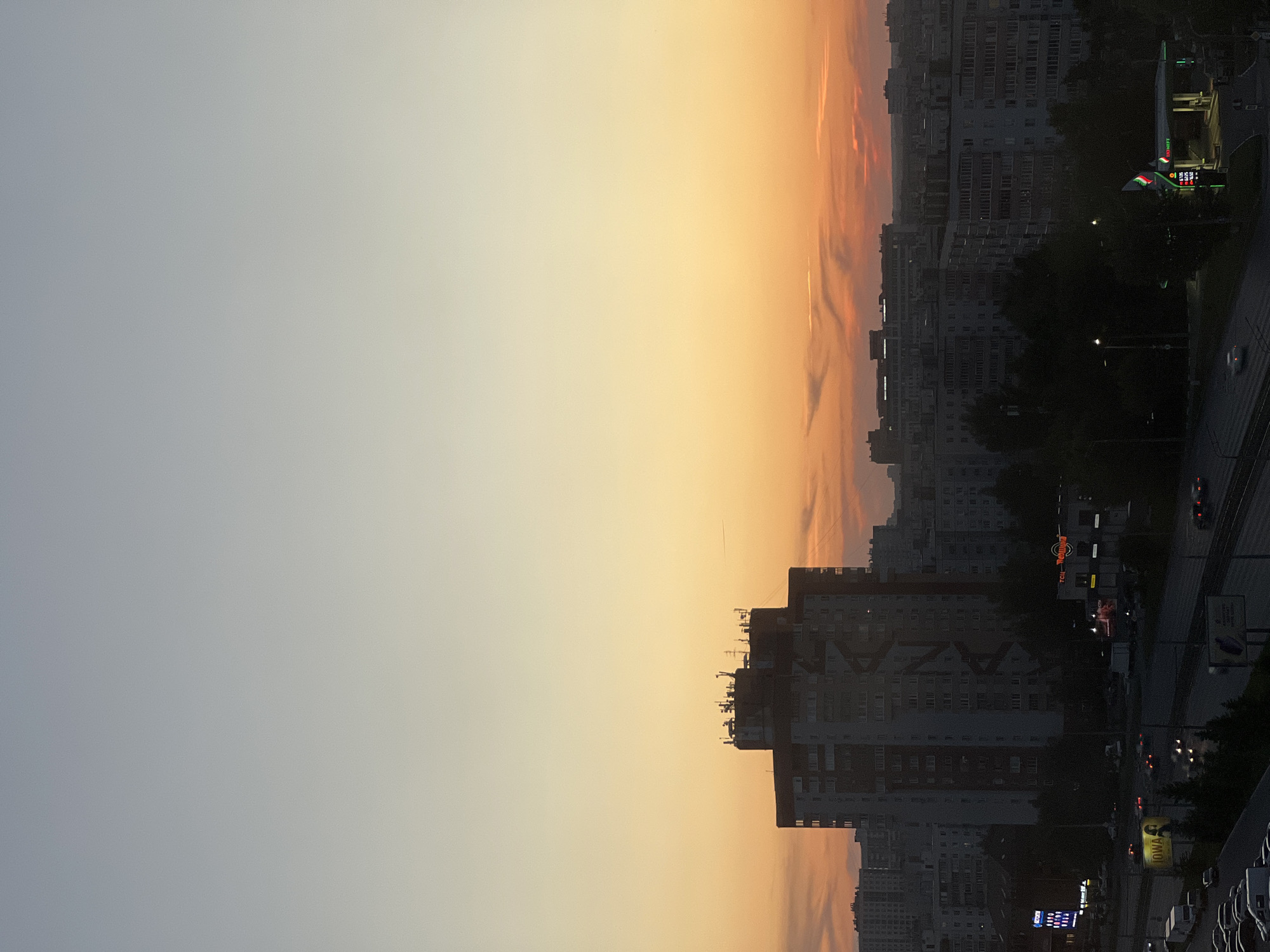 This is a high-angle, full-frame photograph of a city skyline at dusk, presumably in Kazan, Russia, given the presence of an "Ineos" gas station, a Russian company. The sky transitions from a pale grey at the top to a vibrant orange and red gradient near the horizon. Silhouetted against this sunset are numerous multi-story buildings, typical of Soviet-era architecture. In the mid-ground, a prominent, tall, rectangular building stands out, with several antennas and communication equipment mounted on its rooftop. Below, a highway is visible with active traffic, indicated by the headlights and taillights of cars. Some streetlights are also illuminated. Commercial signs, including one with "Ineos" in English and Russian Cyrillic, are visible near the gas station. The overall atmosphere is urban and industrial, captured during the transitional light of sunset.