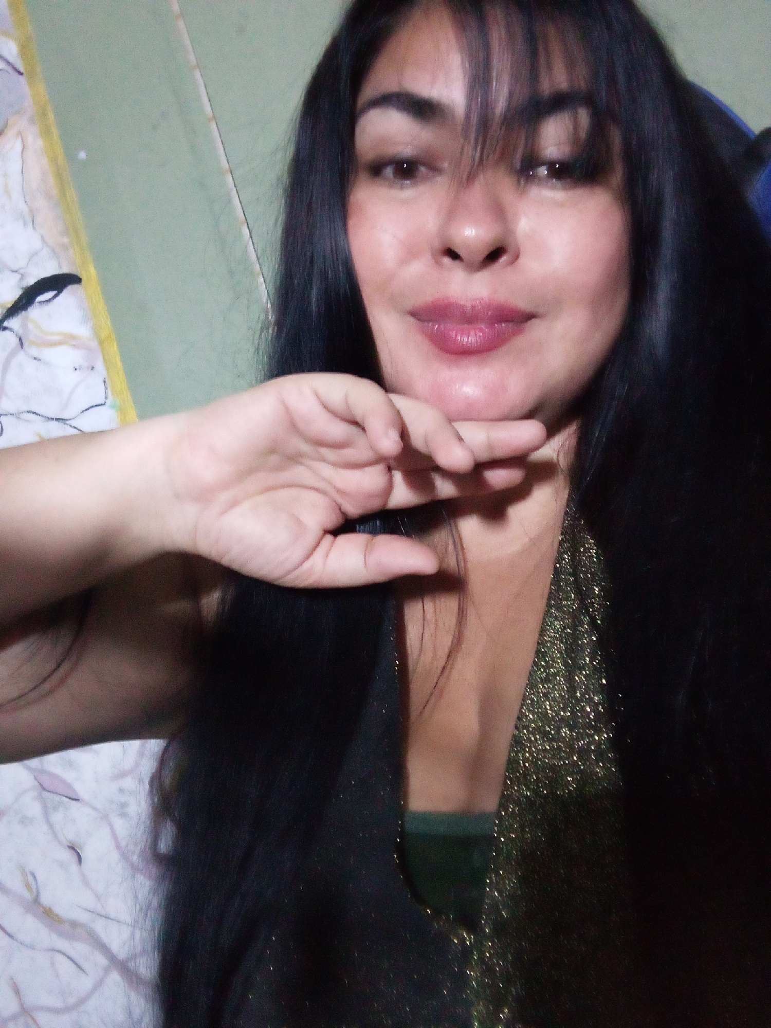 A woman with dark hair and bangs is posing for a selfie. She has her hand to her chin. She is wearing a dark top with a gold sparkly overlay. The background is a light green wall and a patterned sheet. There is no evidence to support the claim that this image was taken in Juan Griego, Venezuela.
