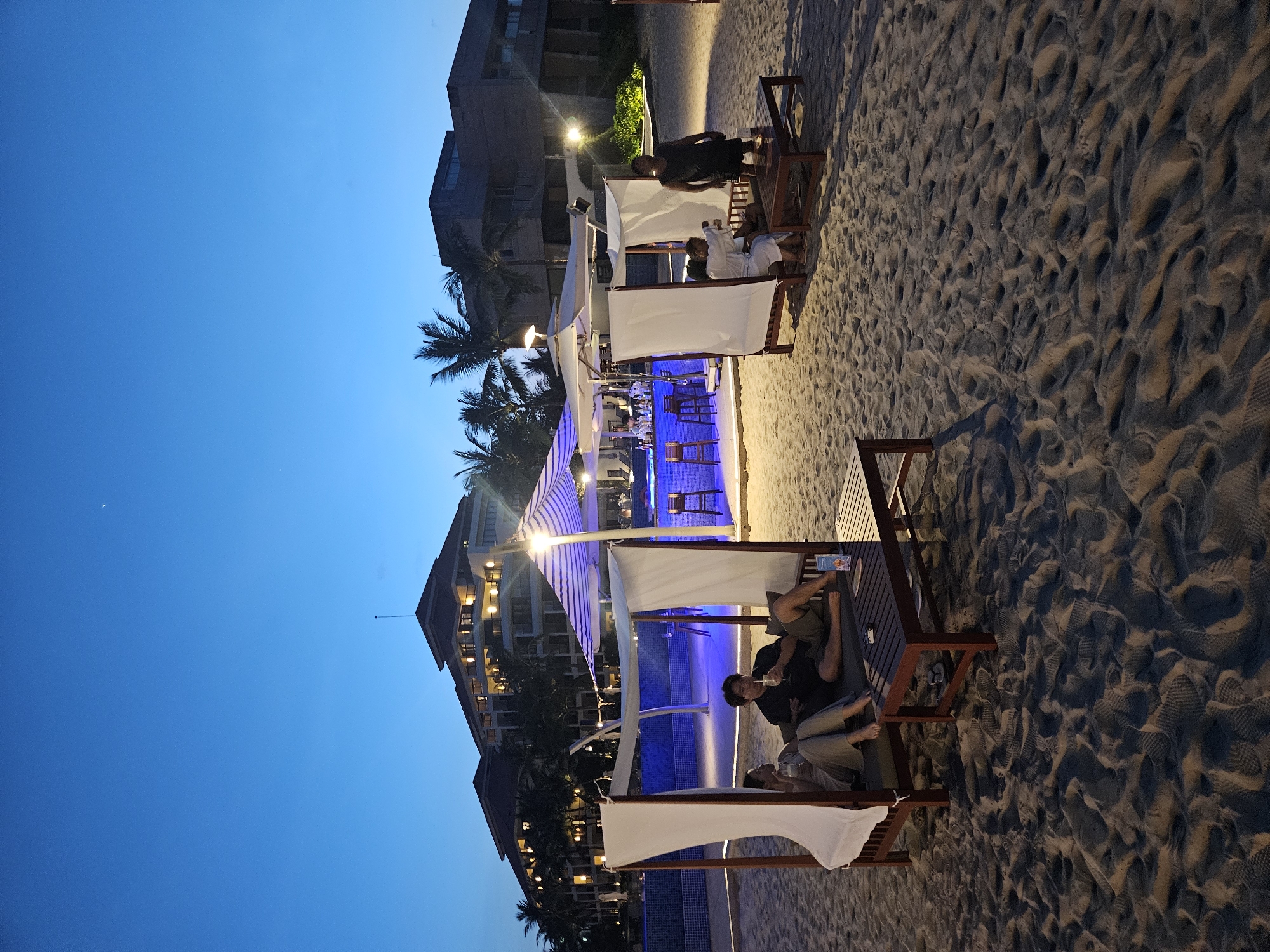 The image depicts an outdoor beach bar area at dusk, likely in Da Nang, Vietnam, as suggested by the presence of palm trees and tropical architecture in the background. In the foreground, two individuals are relaxing on a daybed under a canopy on the sandy beach. One person, dressed in light-colored clothing, is seated and facing away from the camera, while the other, in a dark t-shirt and shorts, is also seated and holding a drink. A low wooden table is placed in front of them.
Further back, another daybed setup features two individuals seated. One is wearing a white robe and appears to be talking, while the other is seated and holding a drink. To the right of this setup, a man stands, wearing a dark tank top and shorts.
Behind the daybeds, a bar area is visible, illuminated by blue lights. Bottles and glasses are arranged on the bar counter, and stools are positioned in front of it. A large umbrella with blue and white stripes provides shade over the bar. In the background, a multi-story hotel or resort building with illuminated windows and balconies is visible, surrounded by palm trees. The sky is a deep twilight blue, indicating the time is shortly after sunset. The sand is textured with footprints and ripples.