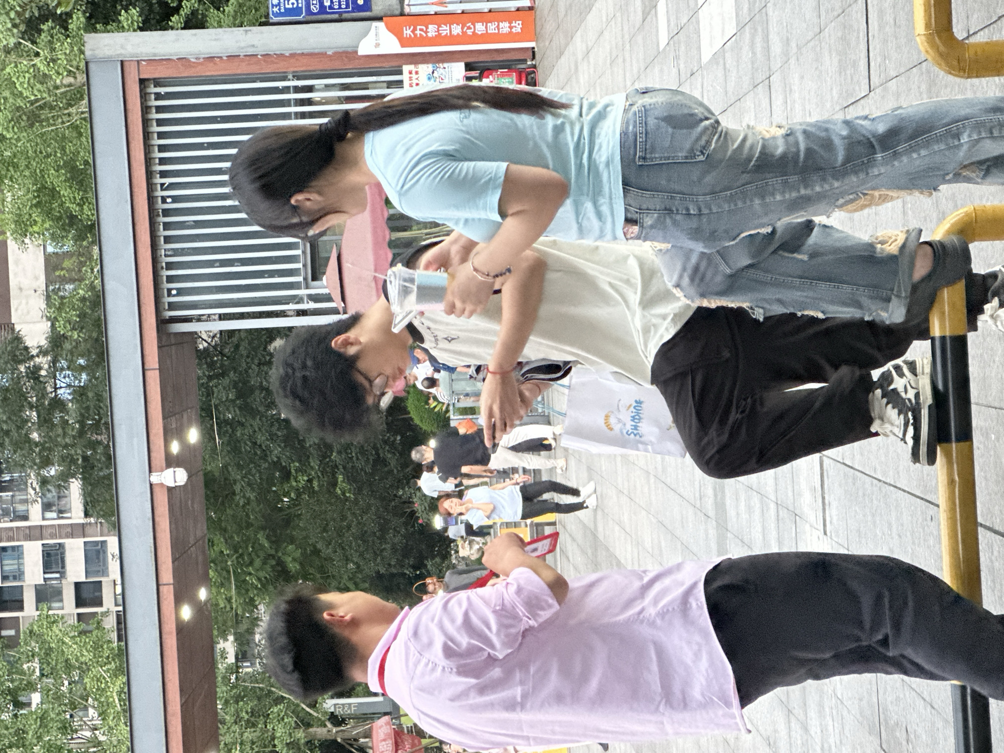 This image depicts a lively outdoor scene, possibly a street or public square, with several people engaged in everyday activities. The overall orientation is vertical, suggesting the photo was taken in portrait mode or rotated.
In the foreground, a young person wearing a light pink collared shirt and black pants is captured from behind, looking towards a group of people further down. To their left and slightly ahead, another young person in a white t-shirt and black pants is interacting with someone holding a clear cup with a straw. Their hair is dark and curly, and they appear to be wearing glasses. Another individual, a young woman with dark hair pulled back in a ponytail, is wearing a light blue t-shirt and distressed denim jeans. She is leaning forward, seemingly observing or participating in the interaction in front of her.
In the background, more people are visible, some walking or standing, suggesting a public gathering or a busy thoroughfare. There are hints of buildings and trees, indicating an urban or semi-urban environment. The presence of a yellow railing and a metal structure with horizontal bars adds to the setting.
Several snippets of Chinese text are visible. One prominent sign reads "天力物业爱心便民驿站" which translates to "Tianli Property Loving Convenience Station," indicating a community service point. Other visible text includes "大学" (University) and "DAXUR". The time of day appears to be daytime, with diffused lighting suggesting it might be overcast or late afternoon. The overall mood seems casual and observational, capturing a moment of social interaction amidst a bustling environment. The location context of Chongqing High-tech Zone suggests a modern, developed area.