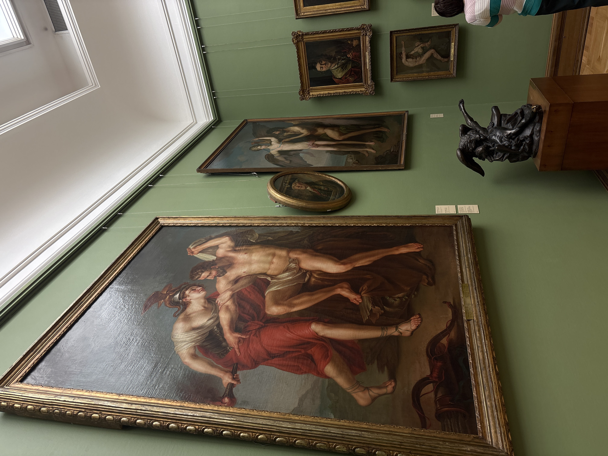 The image depicts a room in the Pushkin State Museum of Fine Arts in Moscow, Russia. On the green-painted wall, various framed paintings are hung, including one large painting depicting two figures. One figure is male, nude and lying across the painting, while the other is female and partially clothed in red, holding a rod-like object. Above this painting, there are several smaller framed paintings, and a round, framed object. To the right of these paintings, a wooden pedestal supports a black sculpture of intertwined figures. The ceiling is white, with decorative trim and recessed lighting.