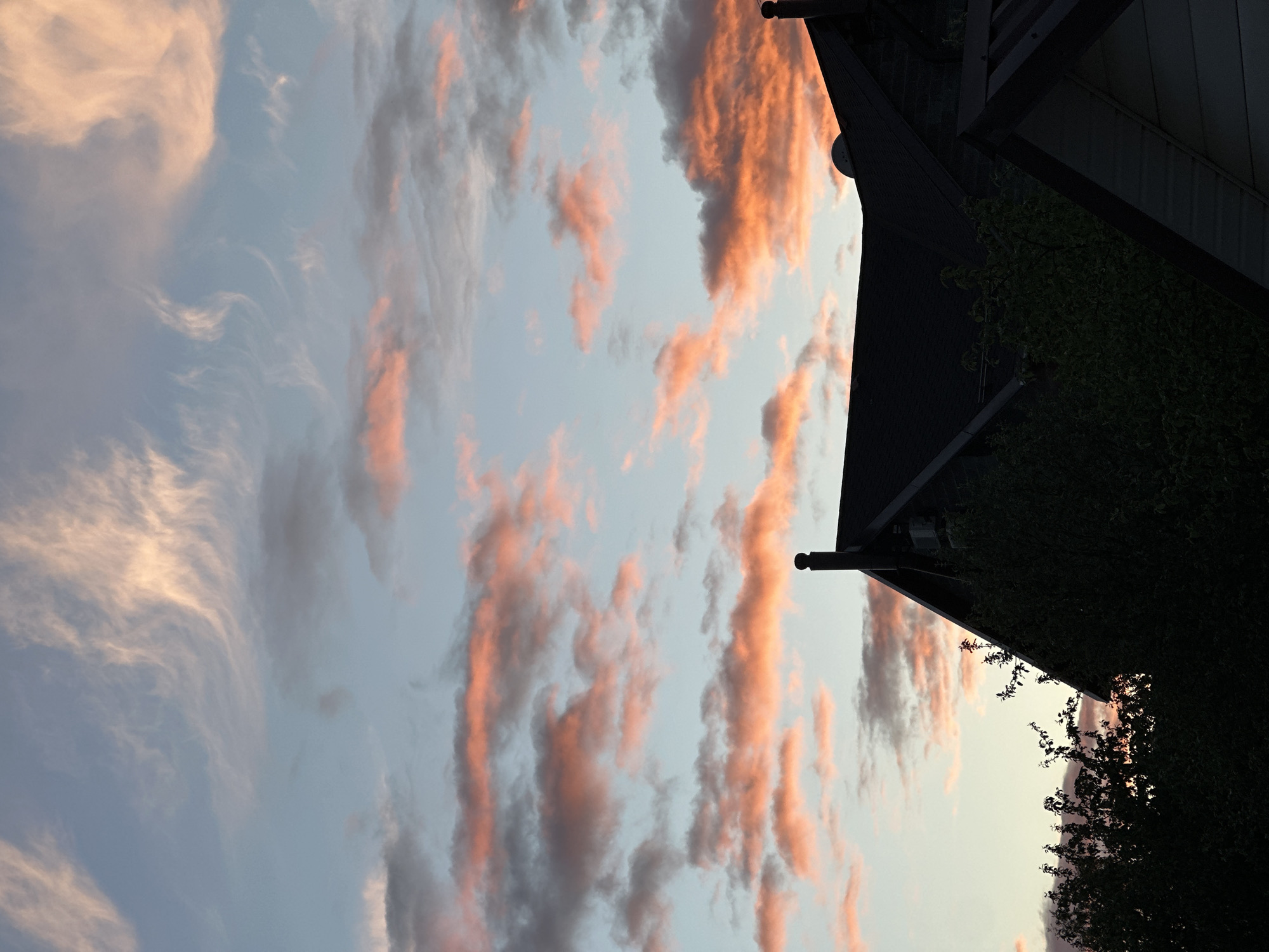 The image depicts a view of the sky at dusk or dawn, with orange-tinted clouds stretched across a pale blue background. The angle suggests the photograph was taken looking upwards, with a portion of a building visible in the upper right quadrant. The building appears to have dark eaves, a gutter system, and is partially obscured by foliage. The overall scene conveys a serene or peaceful atmosphere. The location is not discernible from the visual information.