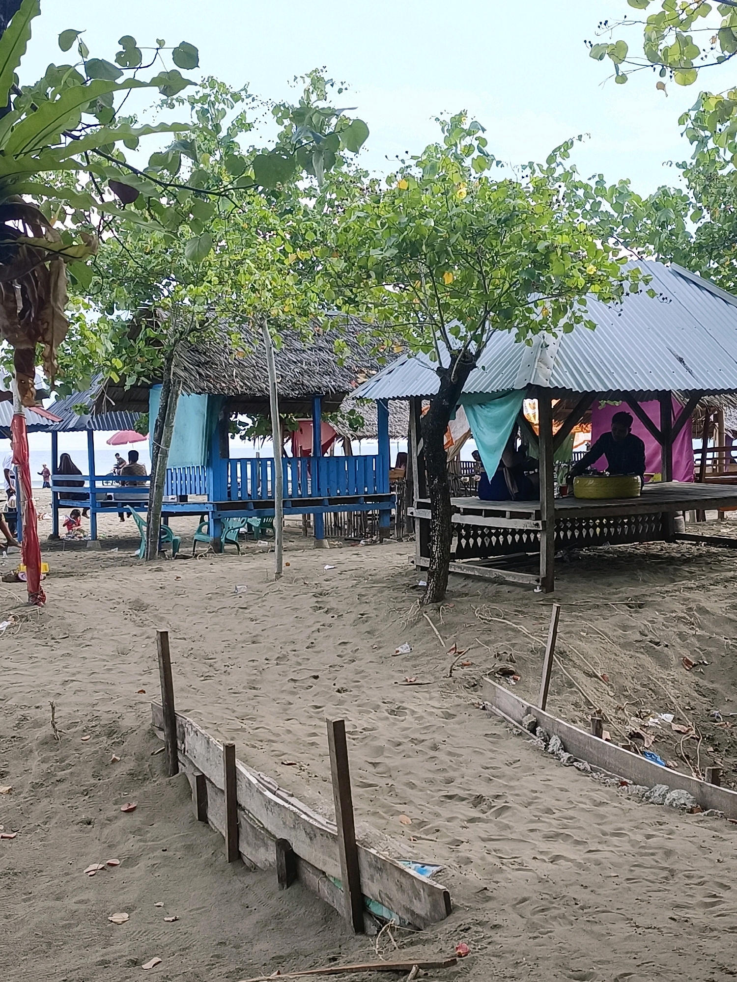 The image depicts a beach scene in Reudeuep, Indonesia. The foreground is dominated by sandy ground with a low, makeshift wooden barrier running diagonally across it. Scattered debris, including leaves and plastic, is visible on the sand.
In the mid-ground, several open-air gazebos or huts are set up on a raised wooden platform. One gazebo, painted blue, has a thatched roof and blue railings. Another, with a corrugated metal roof, is decorated with colorful fabric drapes in turquoise and pink. A person is seated inside this latter gazebo, engaged with a yellow object. Other gazebos with thatched roofs are also present, some with people seated within.
Further back, a body of water, likely the sea, is visible beyond the gazebos. A red umbrella is open near the blue gazebo, suggesting people are seeking shade. The sky is overcast. Trees with green foliage are situated around the gazebos, with some branches extending into the frame at the top. The overall impression is of a casual, perhaps slightly dilapidated, beachside recreational area.