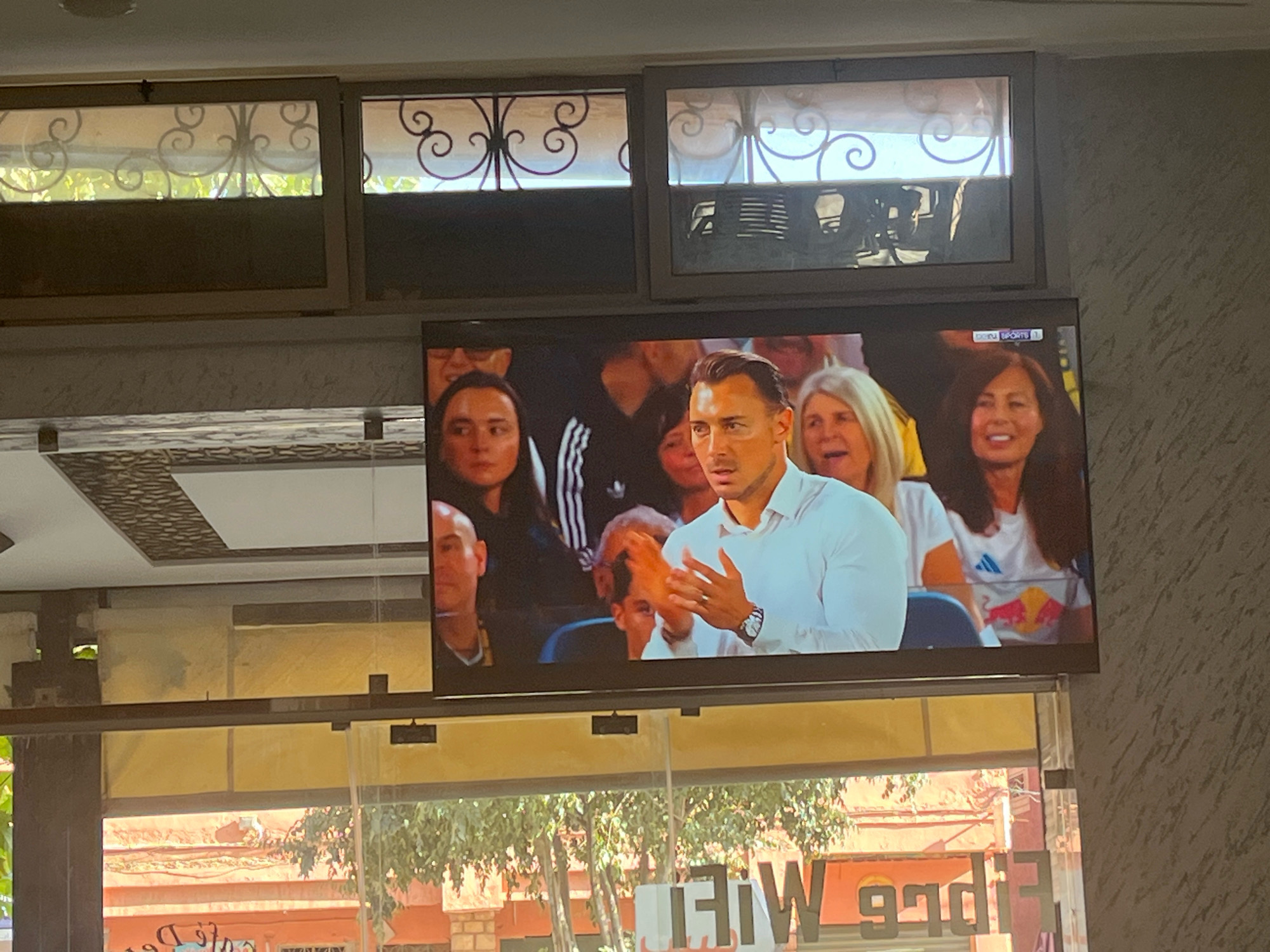 This image shows a television screen displaying a live broadcast of a sports event, likely football, with several people visible. The main subject on the screen is a man in a white shirt clapping his hands. He is surrounded by other spectators. The television is mounted on a wall above a window. Below the window, there is signage with text that appears to be "FiDi Wi-Fi", indicating this is likely a cafe or establishment offering internet service. The scene suggests an indoor setting in Azilal, Morocco, possibly a cafe or restaurant, with a view of the outdoors through the window. The architecture visible in the background through the window suggests a typical Moroccan urban or semi-urban environment.
