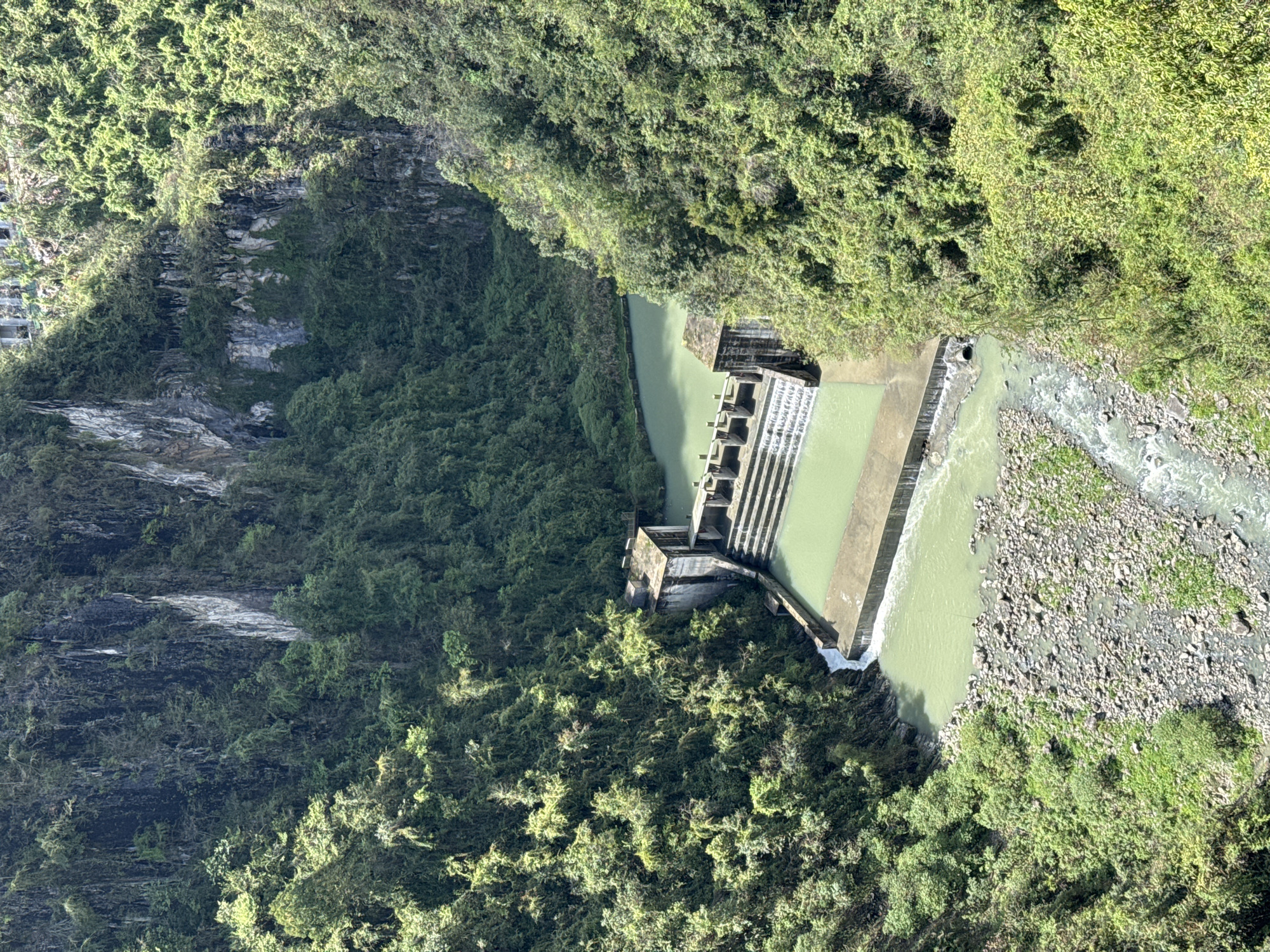 Aerial view of a river and dam surrounded by lush green vegetation in Qianjiang, China. The river's water has a light green tint. A concrete dam structure spans the river. Downstream from the dam, the riverbed is visible, consisting of rocks and sparse vegetation. The surrounding landscape is densely covered with trees and foliage. The overall scene depicts a natural setting with a man-made structure integrated into the environment.