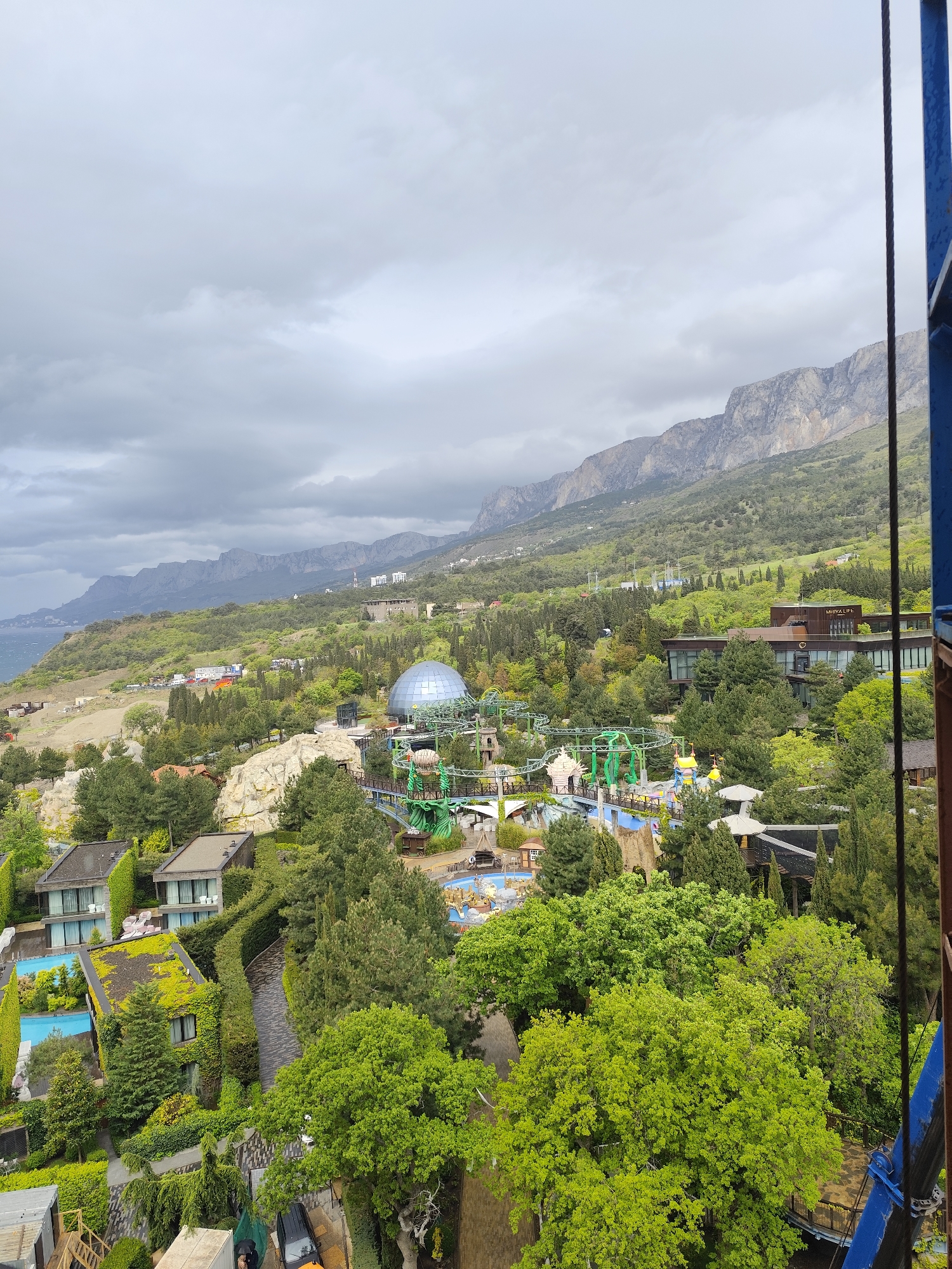 The image shows a view of a landscape with a combination of natural and man-made features. A range of mountains is visible in the background under a cloudy sky. The middle ground is filled with lush greenery including trees and landscaped areas. In this area, there is a structure with a domed roof. A water park or amusement park is evident with visible rides. Residential or resort buildings are also seen, some with green roofs. The image appears to be taken from an elevated position, possibly from a tall structure or a high vantage point. A partial view of a blue structure is visible on the right side of the frame.