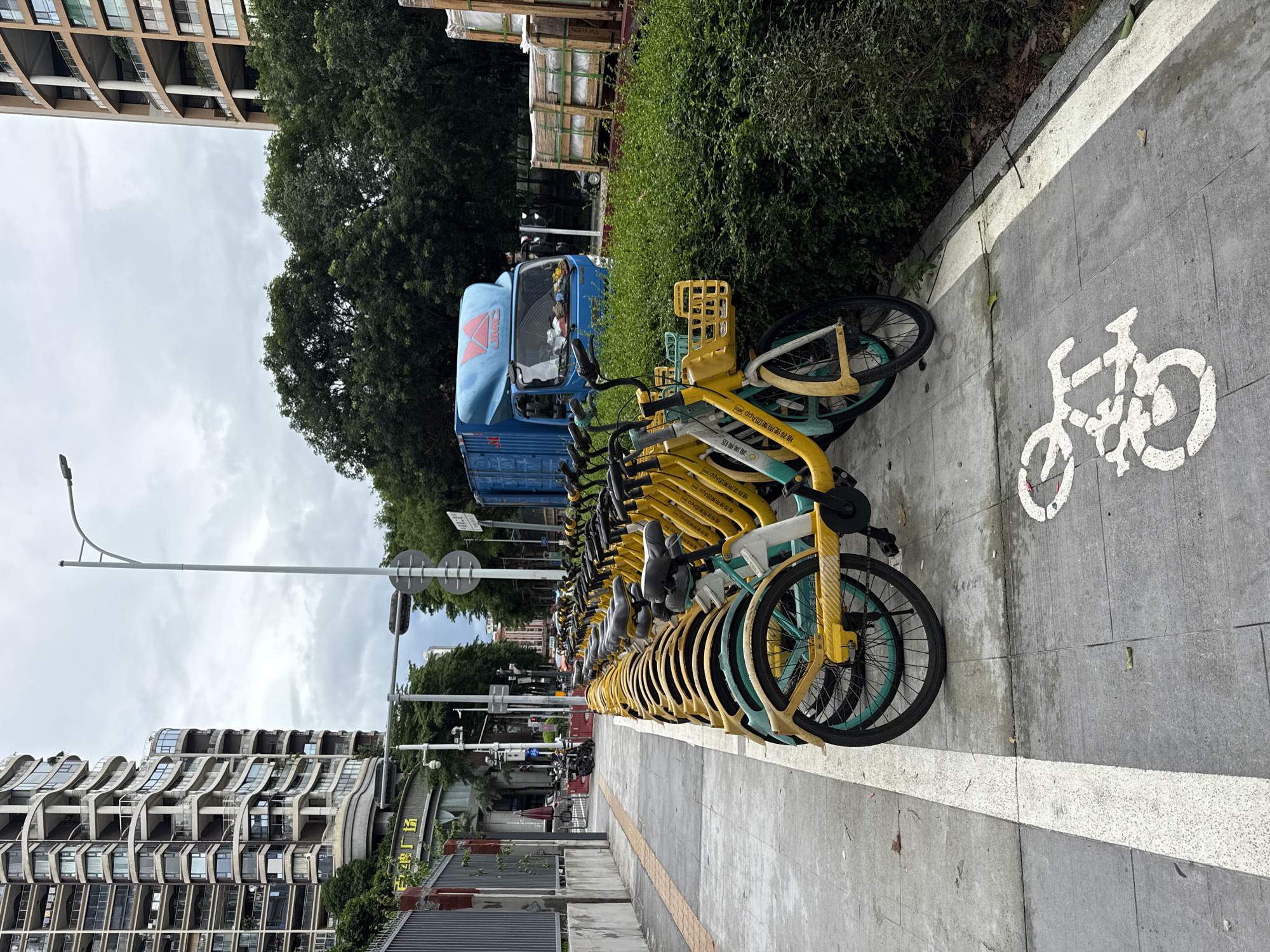 This image captures a row of shared bicycles parked on a sidewalk in Nanshan District, China. The bicycles are predominantly yellow with teal accents and have baskets on the front. They are lined up in a designated parking area marked by a white bicycle symbol painted on the grey pavement. In the background, a tall, modern residential building with numerous balconies dominates the left side of the frame. To the right, a blue delivery truck is parked near stacks of wooden crates containing what appear to be glass panels. Lush green trees and bushes line the right side of the scene, bordering a green space. The sky is overcast, suggesting a cloudy day. A street light stands tall on the left, and traffic signals are visible in the distance.