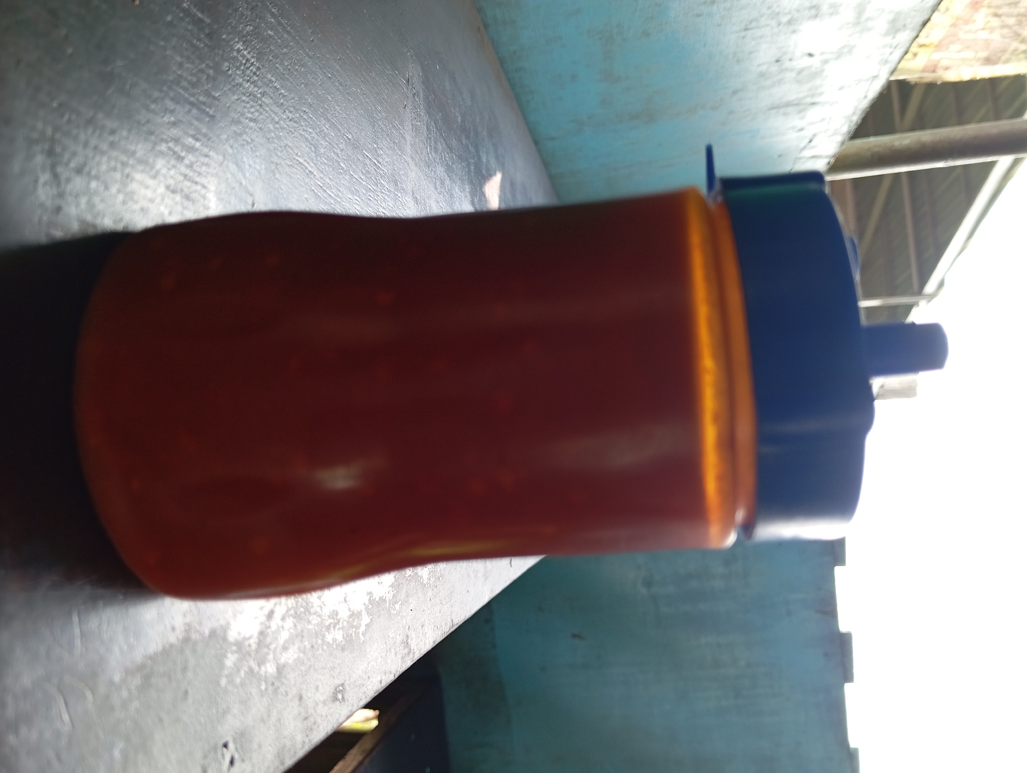 A reddish-orange liquid fills a clear plastic bottle with a blue cap. The bottle is positioned on a dark, textured surface that appears to be a table or ledge. In the background, a light blue wall is visible, with a structure of metal poles and a corrugated roof above it. The scene is outdoors, as indicated by the bright sky and sunlight casting shadows. This image was taken in Lemahbang, Indonesia.