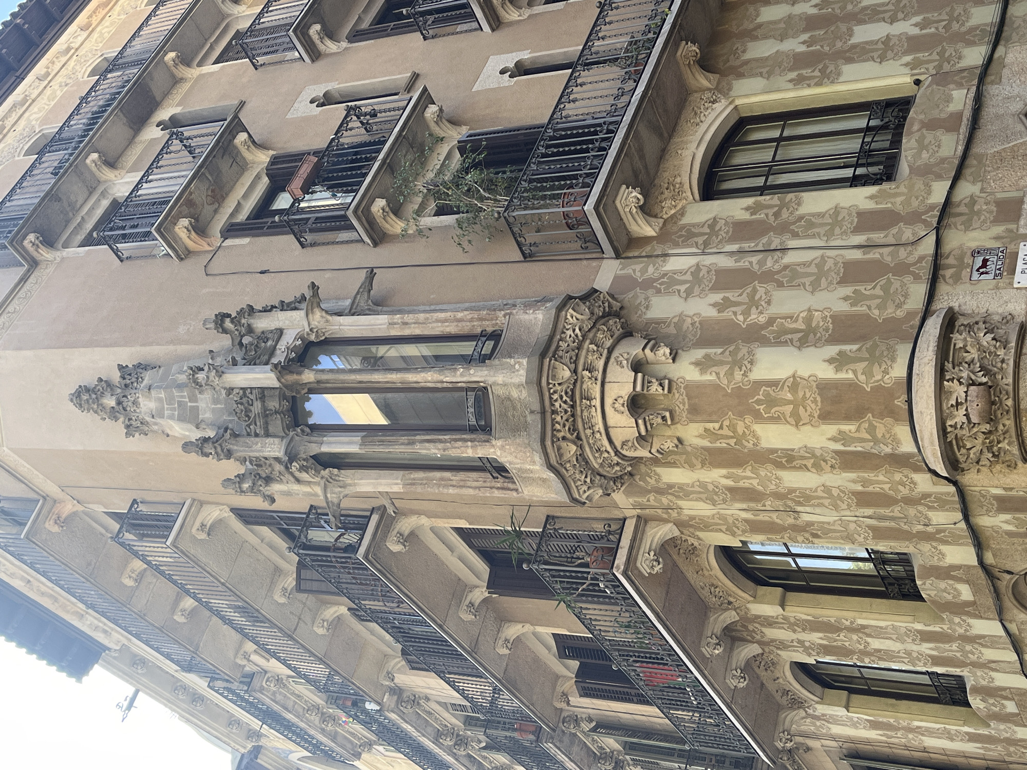 A close-up, upward-angled view captures the ornate facade of a historic building in Barcelona, featuring distinctive Modernisme (Catalan Art Nouveau) architectural elements. The image is rotated, presenting the building's vertical face horizontally.
The building's surface is largely covered in a patterned sgraffito design of repeated floral or leafy motifs in shades of beige and light brown. Numerous rectangular balconies with dark, intricate wrought-iron railings extend from the facade. Many windows are visible, some with internal blinds, and a notable one on the patterned section is horizontally oriented and features additional dark grates.
A prominent feature is a multi-story, highly decorated oriel window or bay window positioned centrally. This structure is richly adorned with sculptural stonework, including gothic-inspired elements, finials, and elaborate carvings at its base and sides. Small potted plants and greenery can be seen on several balconies, including a larger plant protruding from one of the upper balconies.
Electrical cables run vertically along the patterned facade. On the right side, a small red and white sign is partially visible, displaying the text "SALIDA" and "PLAÇA," with an illustration of an animal (possibly a horse or bull) below "SALIDA." The scene is brightly lit by natural daylight, suggesting a clear day. No people are visible.