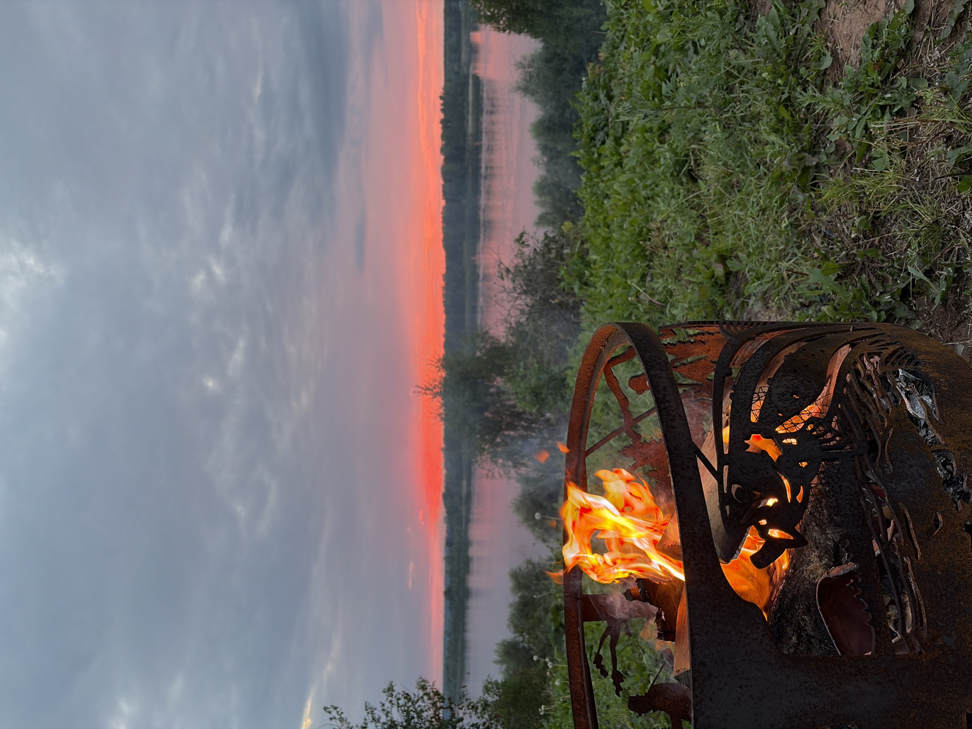 A sunset over a wide river is visible in the background, with a bright orange-pink band of light on the horizon. In the foreground, a rusty metal fire pit contains a lively fire with visible flames and burning logs. The fire pit has intricate cutouts. Green grass and vegetation surround the fire pit, suggesting an outdoor setting. The sky is filled with clouds, tinged with gray. The location is identified as Olkhovka, Russia.